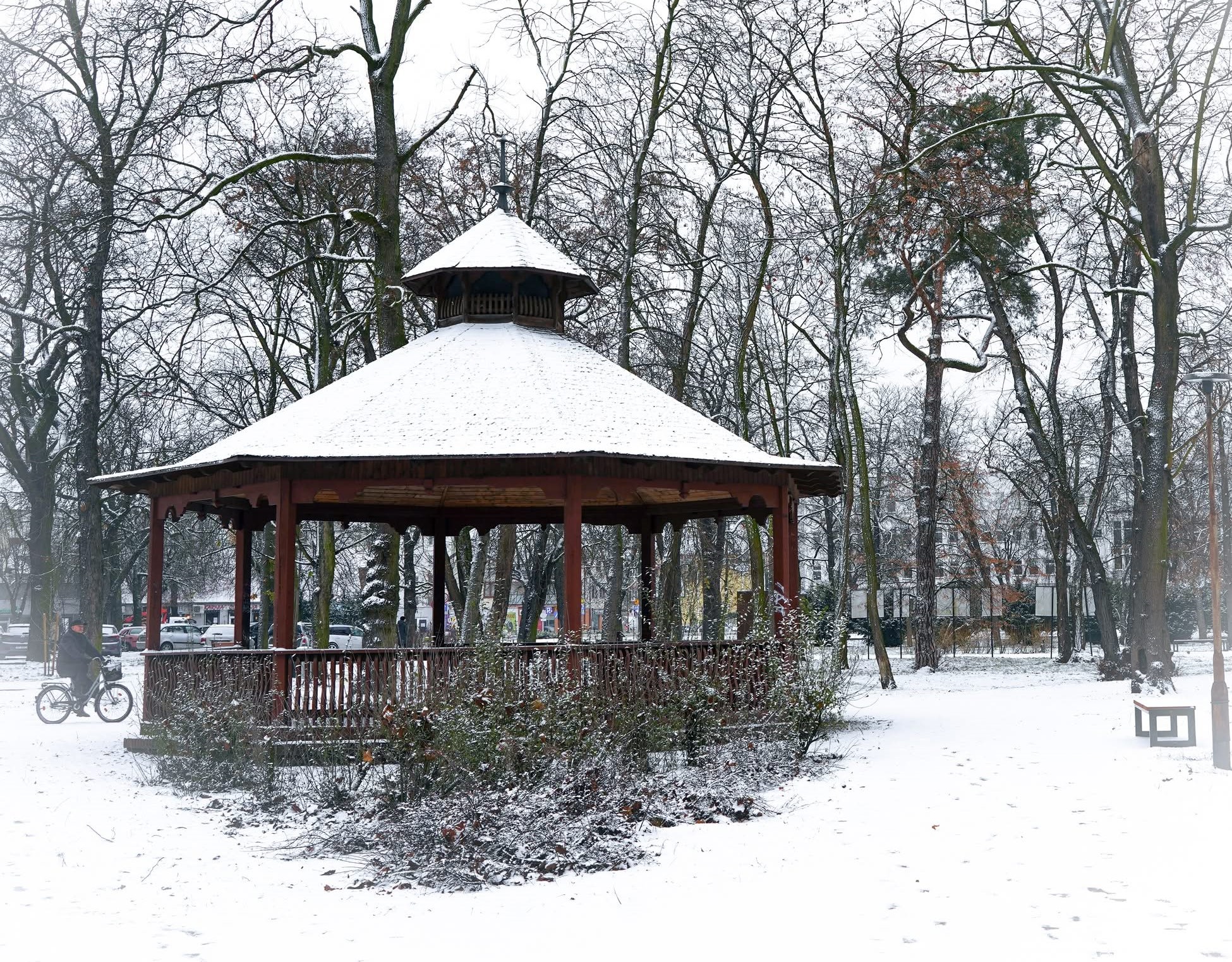 Zdjęcie zrobione w zimowej scenerii na którym widać altanę w parku otoczoną drzewami pokrytymi śniegiem. W tle z lewej strony widać przejeżdżającego rowerzystę.