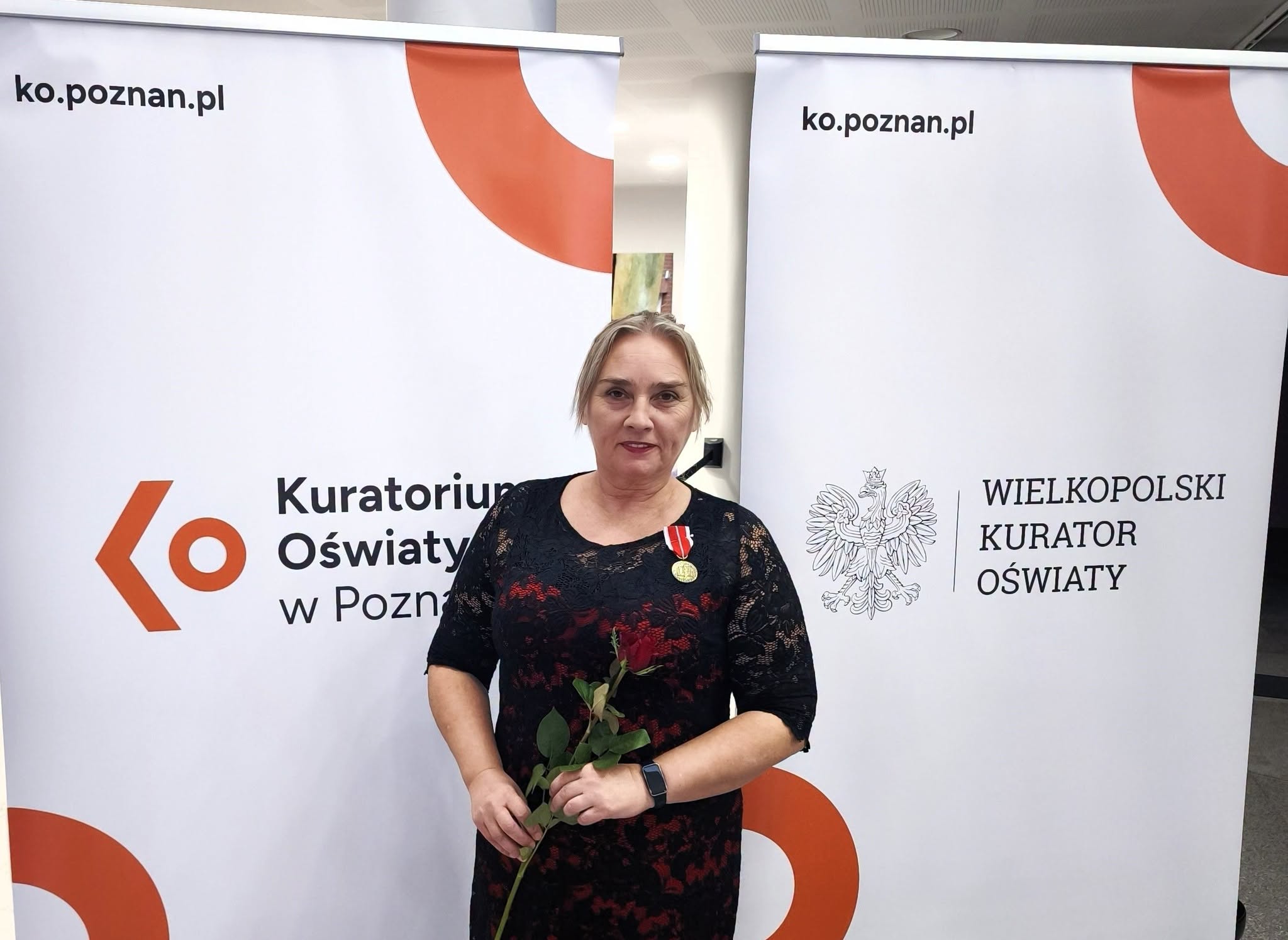 Kobieta w czarnej koronkowej sukience z czerwonymi wstawkami trzyma czerwoną różę. Do sukienki ma przypięty medal na czerwono-białej wstążce. Stoi na tle banerów Kuratorium Oświaty w Poznaniu i Wielkopolskiego Kuratora Oświaty.
