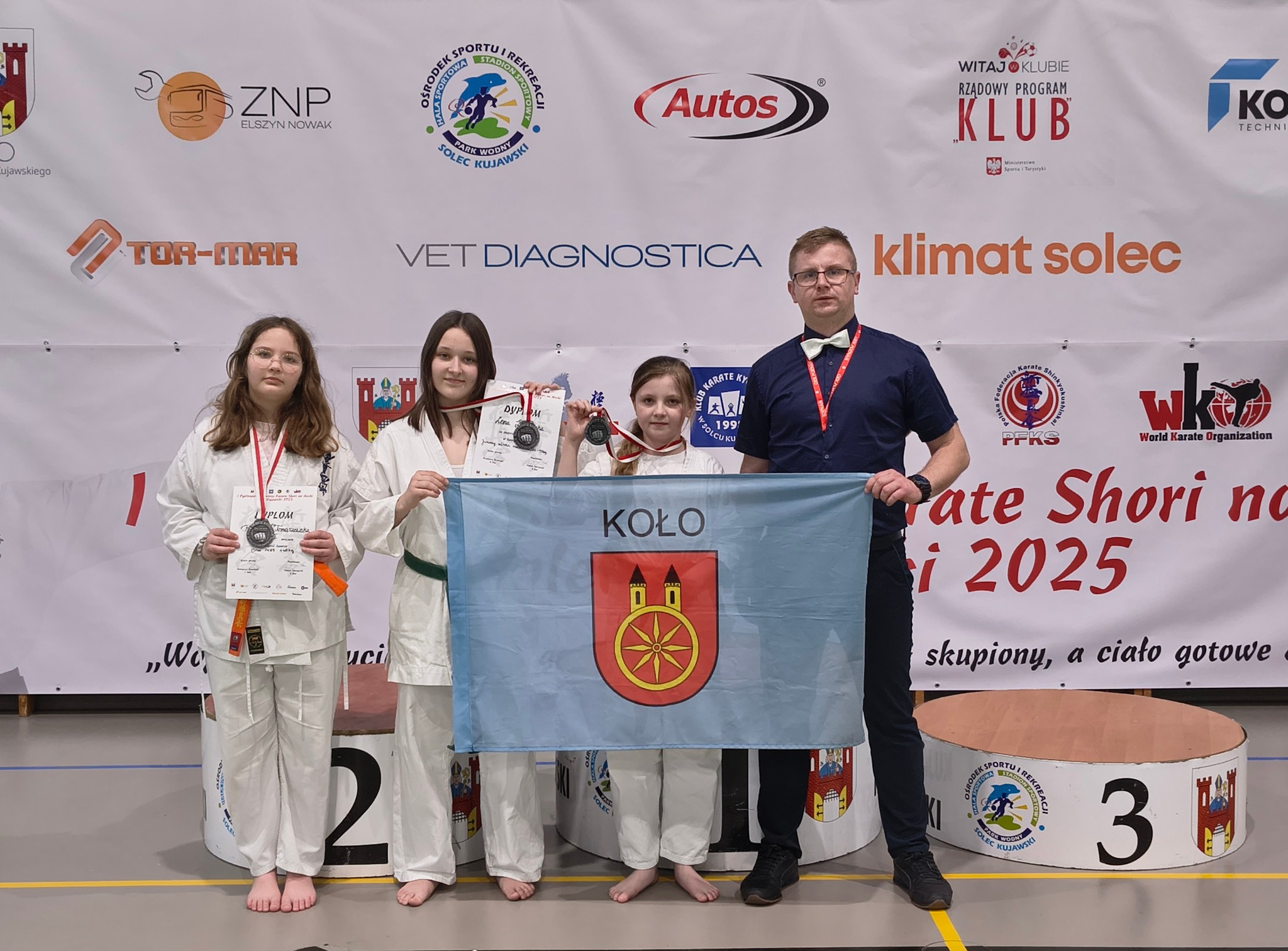 Na zdjęciu widać trzy młode zawodniczki w strojach karate, które stoją przed podium wraz z opiekunem. Zawodniczki trzymają w dłoniach dyplomy oraz mają zawieszone na szyi medale. Jedna zawodniczka oraz mężczyzna trzymają flagę miasta Koła.