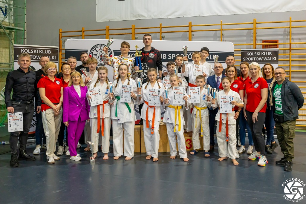 Na zdjęciu widać grupę młodych zawodników w strojach do karate, trzymających puchary i dyplomy, co wskazuje na zakończenie turnieju. W tle widoczne są banery z nazwą Kolski Klub Sportów Walki, a za zawodnikami stoją trenerzy i organizatorzy 