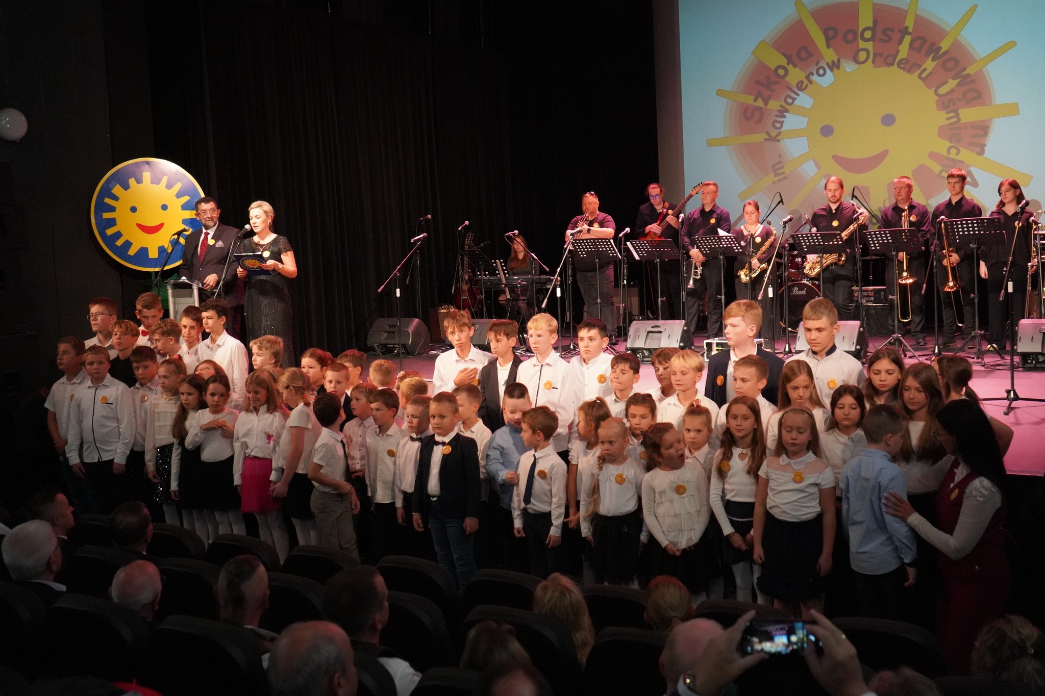 Na zdjęciu widzimy grupę dzieci stojących na scenie w eleganckich ubraniach, podczas występu szkolnego. W tle widać zespół muzyczny oraz logo szkoły z uśmiechniętym słońcem. Na scenie znajduje się również para dorosłych, prowadzących wydarzenie.