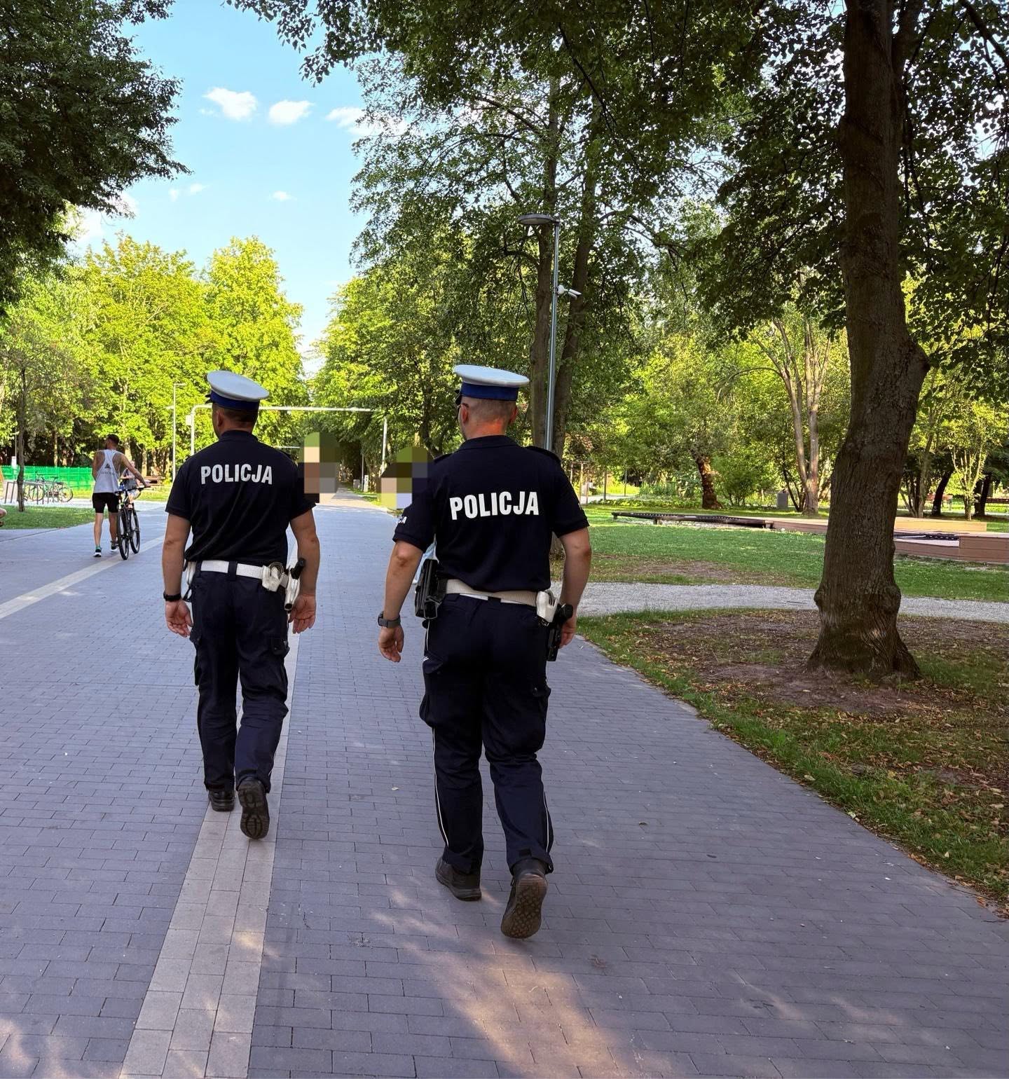 Dwóch policjantów w granatowych koszulkach z napisem POLICJA i białych czapkach idzie, oddalając się od obiektywu, alejką z kostki brukowej przez zielony park. Po lewej mężczyzna prowadzi rower, w tle wysokie drzewa i trawniki.