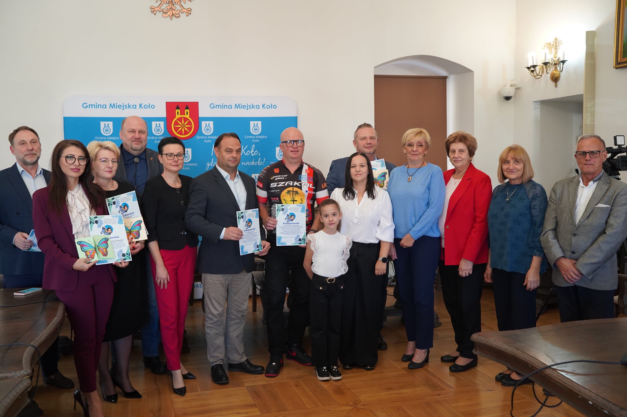 Grupa ludzi pozuje do wspólnego zdjęcia w sali sesyjnej Ratusza, trzymając w dłoniach pamiątkowe dyplomy. W tle widać ściankę promocyjną miasta Koła z napisem MIASTO KOŁO GDZIE ZABYTKI NAOKOŁO.