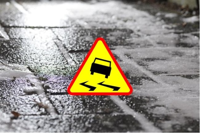 To zdjęcie śliskiego, oblodzonego chodnika z brukowej nawierzchni. Na pierwszym planie żółty znak ostrzegawczy z symbolem samochodu wpadającego w poślizg. Grafika zwraca uwagę na zagrożenie związane z gołoledzią.