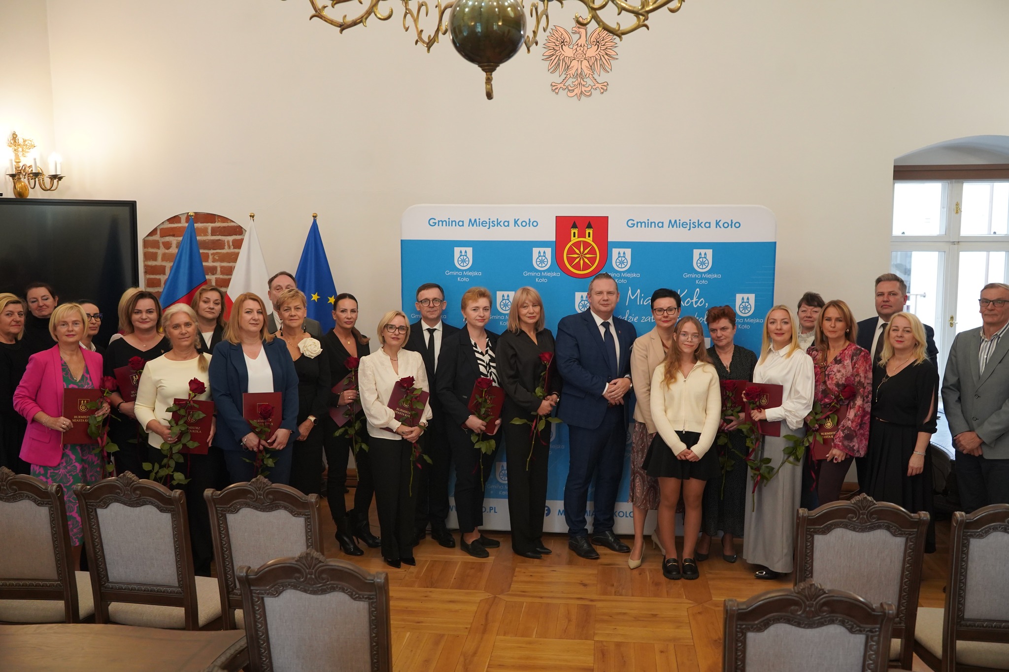 Na zdjęciu widać grupę osób stojących wspólnie w sali urzędowej, trzymających czerwone róże i dyplomy. W tle znajduje się ścianka z herbem miasta Koło oraz flagi Polski, UE. Jest to uroczystość wręczenia nagród.