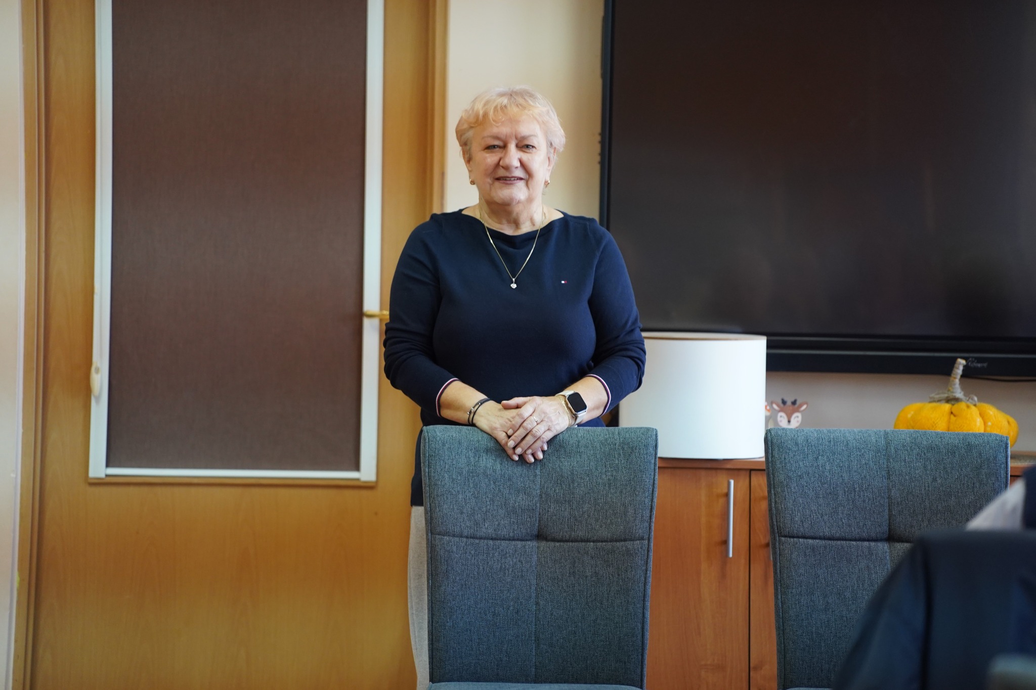 Na zdjęciu widać uśmiechniętą kobietę stojącą w pomieszczeniu, opierającą dłonie na oparciu krzesła; w tle znajduje się duży ekran na ścianie, drzwi oraz komoda, na której stoi ozdobna żółta dynia.