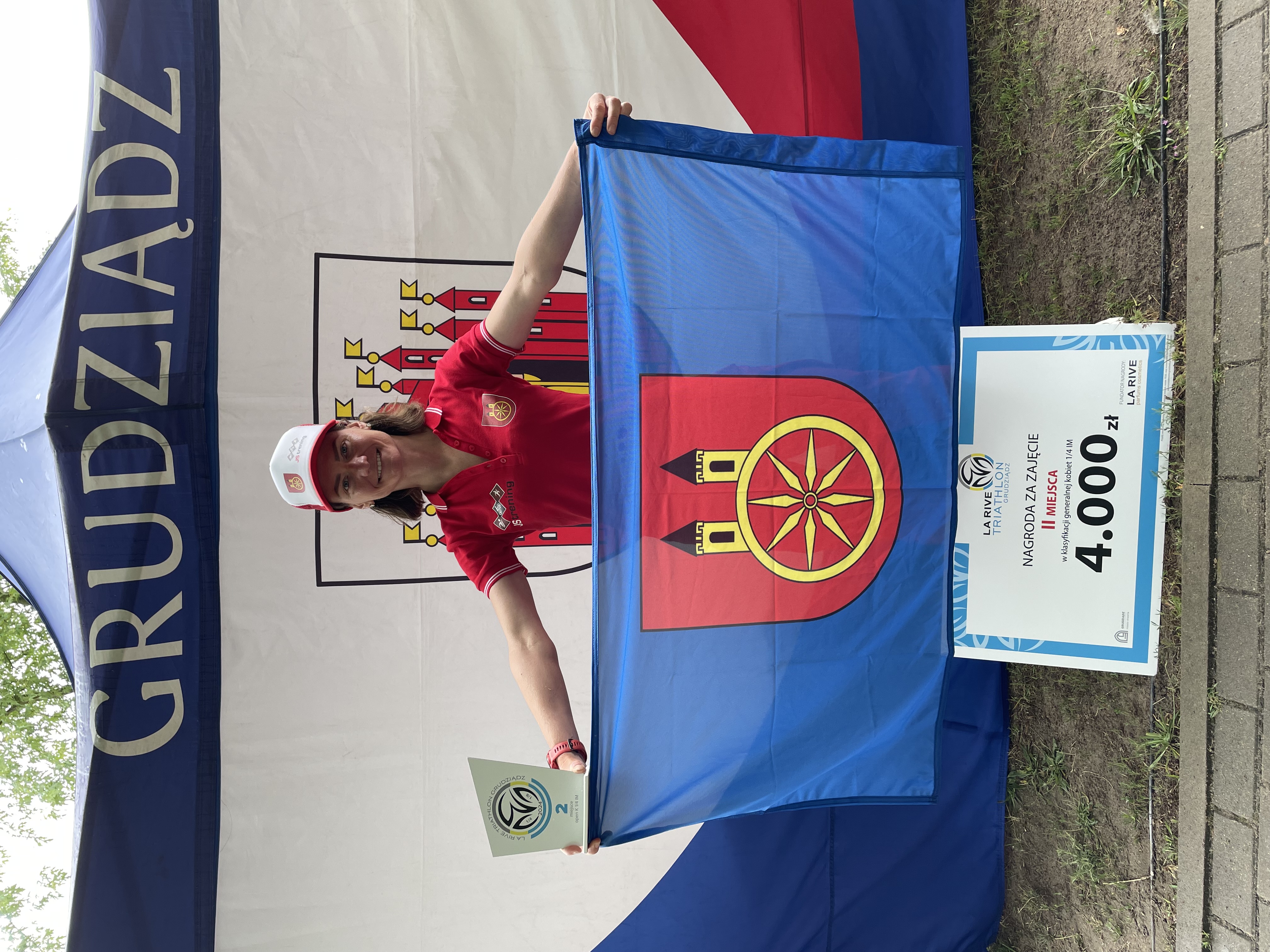 Zdjęcie przedstawia kobietę trzymającą w dłoniach flagę miasta Koła, na dole dyplom