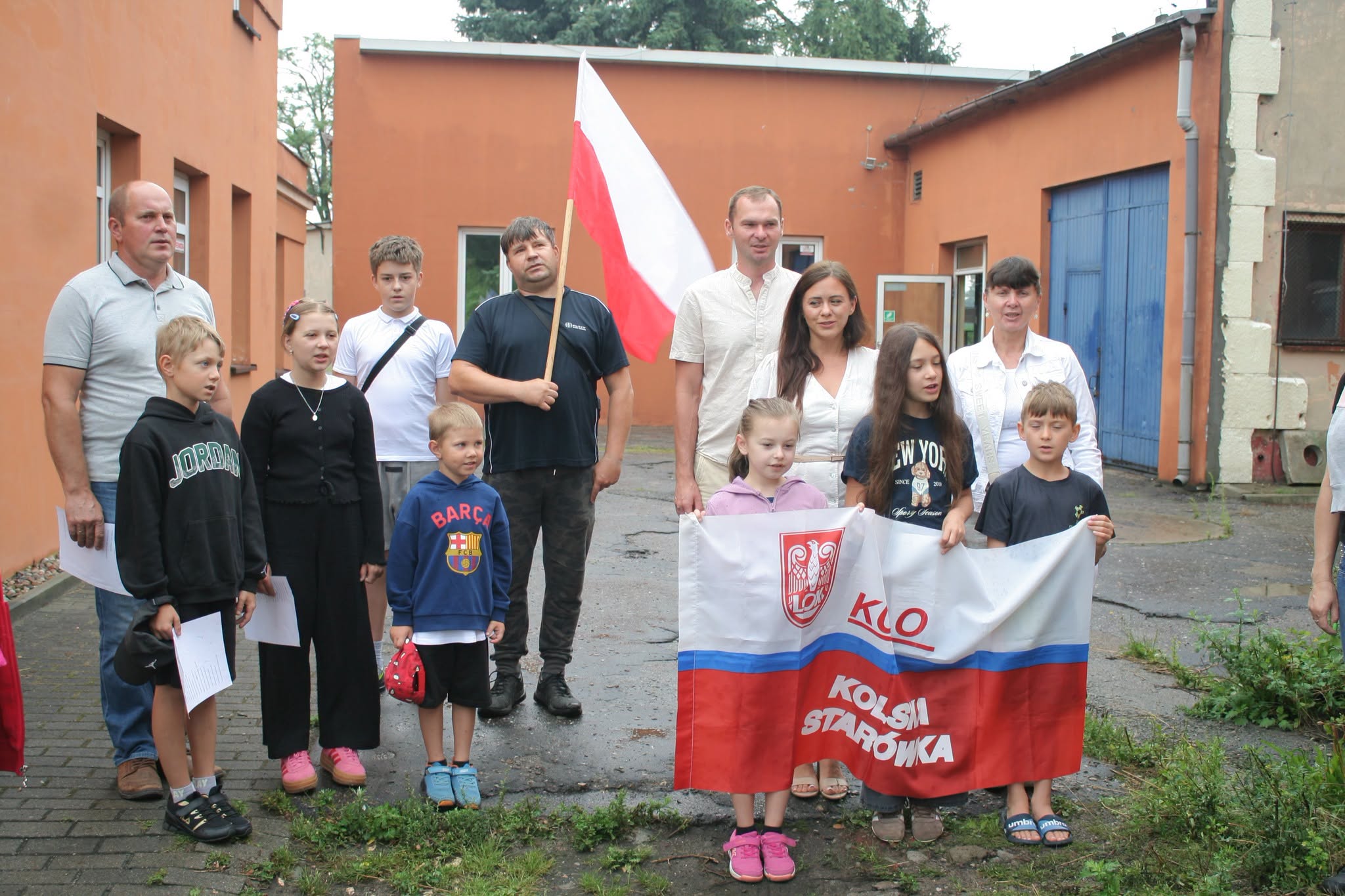 Grupa kilkunastu osób, dorosłych i dzieci, pozuje na podwórzu przy pomarańczowym budynku z niebieską bramą. Mężczyzna w środku trzyma na drzewcu biało-czerwoną flagę Polski, a dzieci z prawej rozwijają biało-czerwony baner z godłem i napisem LOK Kols