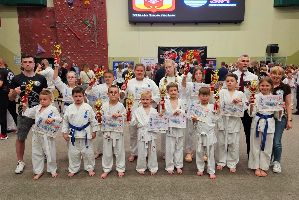 Grupowe zdjęcie dzieci w białych kimonach karate z niebieskimi pasami, trzymających złote puchary i dyplomy. Obok nich dwoje dorosłych opiekunów. W tle hala sportowa z napisem Miasto Inowrocław. Pamiątkowe foto z zawodów.