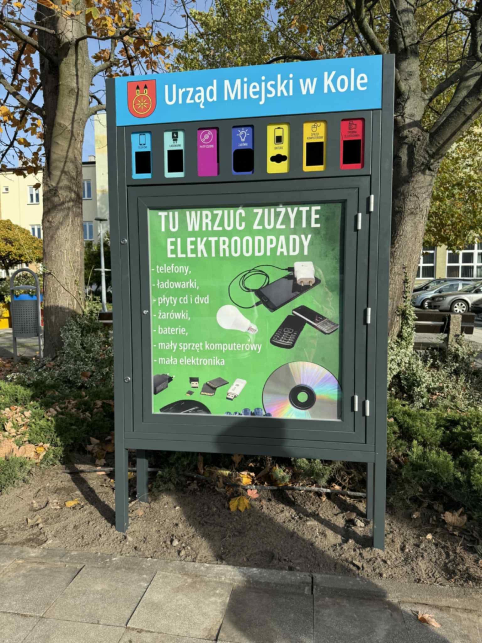 Na zdjęciu widać pojemnik na elektrośmieci przy chodniku, z tyłu dwa drzewa, w tle mały zieleniec, kosz na śmieci, budynki i samochody.