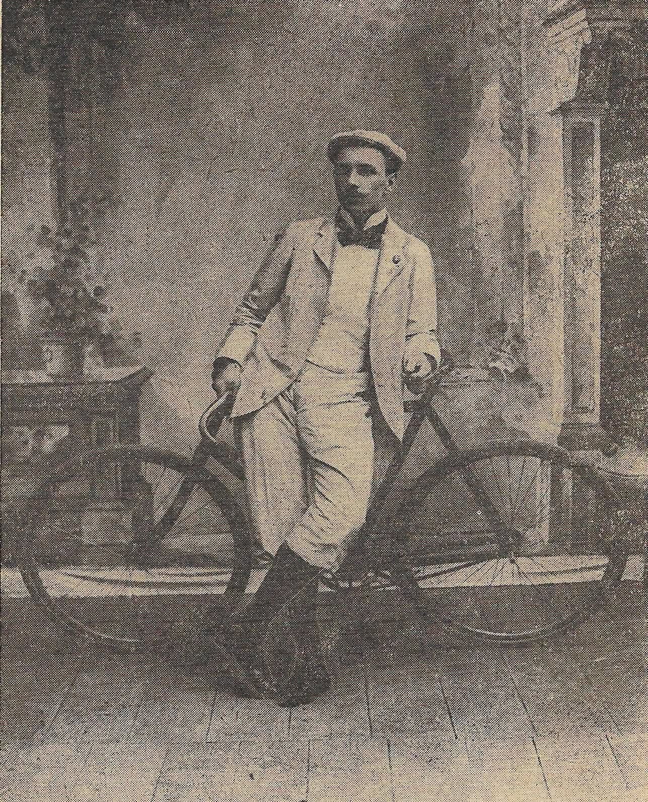 Czarno-białe historyczne zdjęcie lub rycina przedstawiająca mężczyznę w jasnym stroju z XIX wieku, czapce i muszce, opierającego się o rower. Drewniana podłoga, architektoniczne tło.