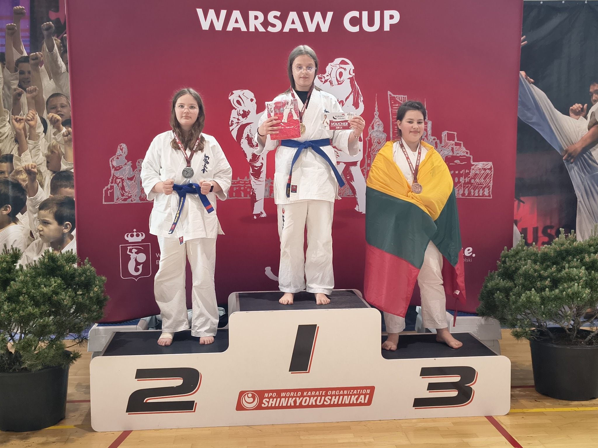 Zdjęcie przedstawia trzy młode zawodniczki karate stojące na podium turnieju Warsaw Cup. Zajęły odpowiednio pierwsze, drugie i trzecie miejsce. Środkowa zawodniczka trzyma puchar i voucher, a trzecia z prawej ma na sobie flagę Litwy.