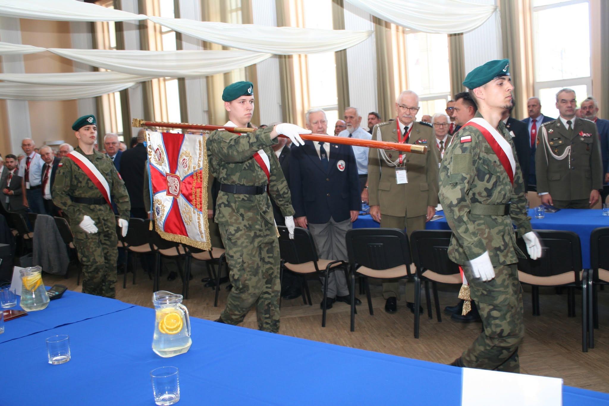 Poczet sztandarowy złożony z trzech uczniów w mundurach polowych i zielonych beretach wnosi sztandar wojskowy podczas uroczystości. W tle stoją oficerowie i goście cywilni w sali bankietowej.