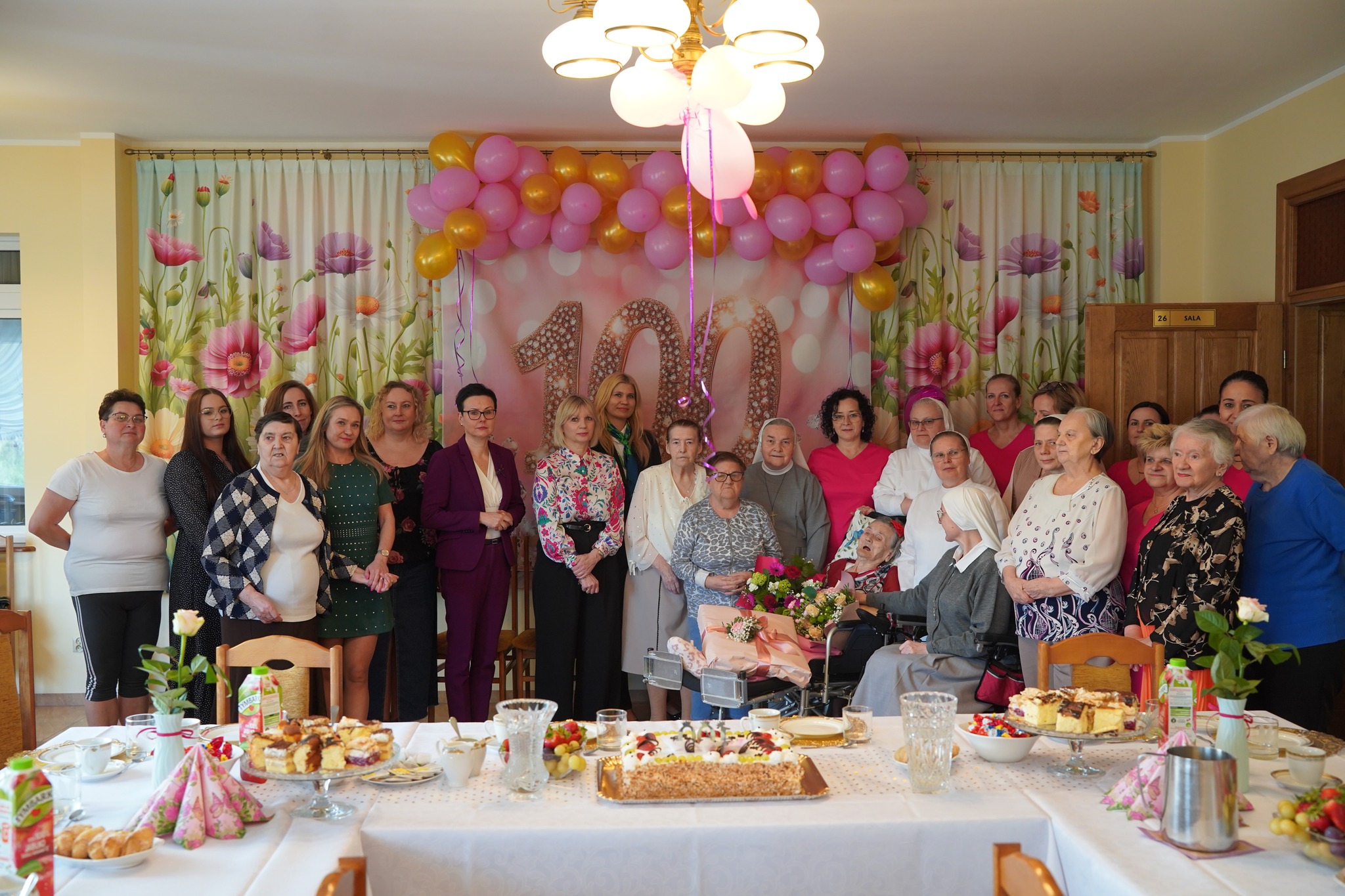 Zdjęcie przedstawia grupę osób świętujących 10o. urodziny starszej kobiety w ozdobionej sali. W tle widoczna jest liczba 100 oraz balony, a na stole znajdują się ciasta i napoje.