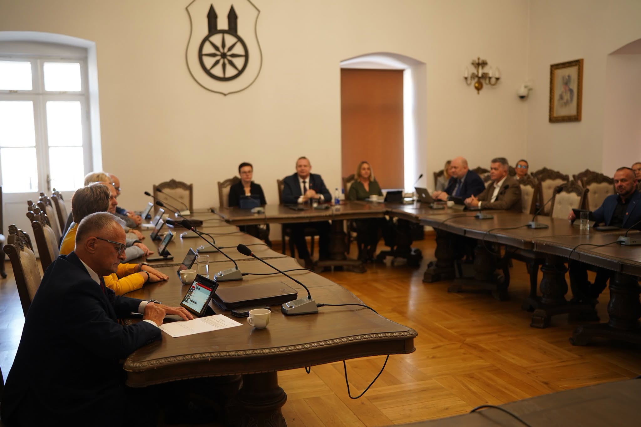 Sala obrad z herbem Koła na ścianie. Radni i goście siedzą przy drewnianych stołach ustawionych w podkowę, z mikrofonami i tabletami. Na pierwszym planie mężczyzna w okularach czyta dokument.