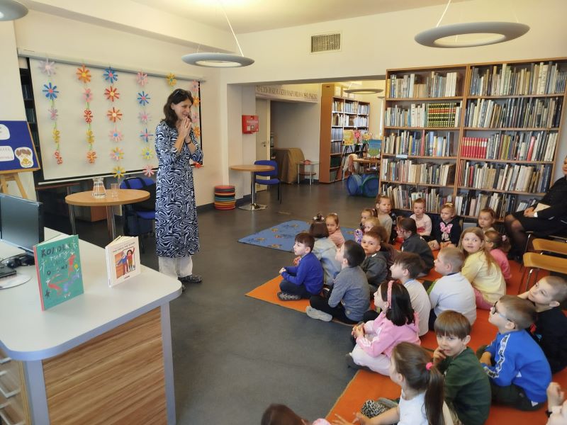 Na zdjęciu stoi autorka i pisarka podczas spotkania w czytelni biblioteki, zwracając się do siedzących na podłodze dzieci, a w tle widać półki z książkami oraz ekran wiszący na ścianie ozdobiony papierowymi kwiatami.