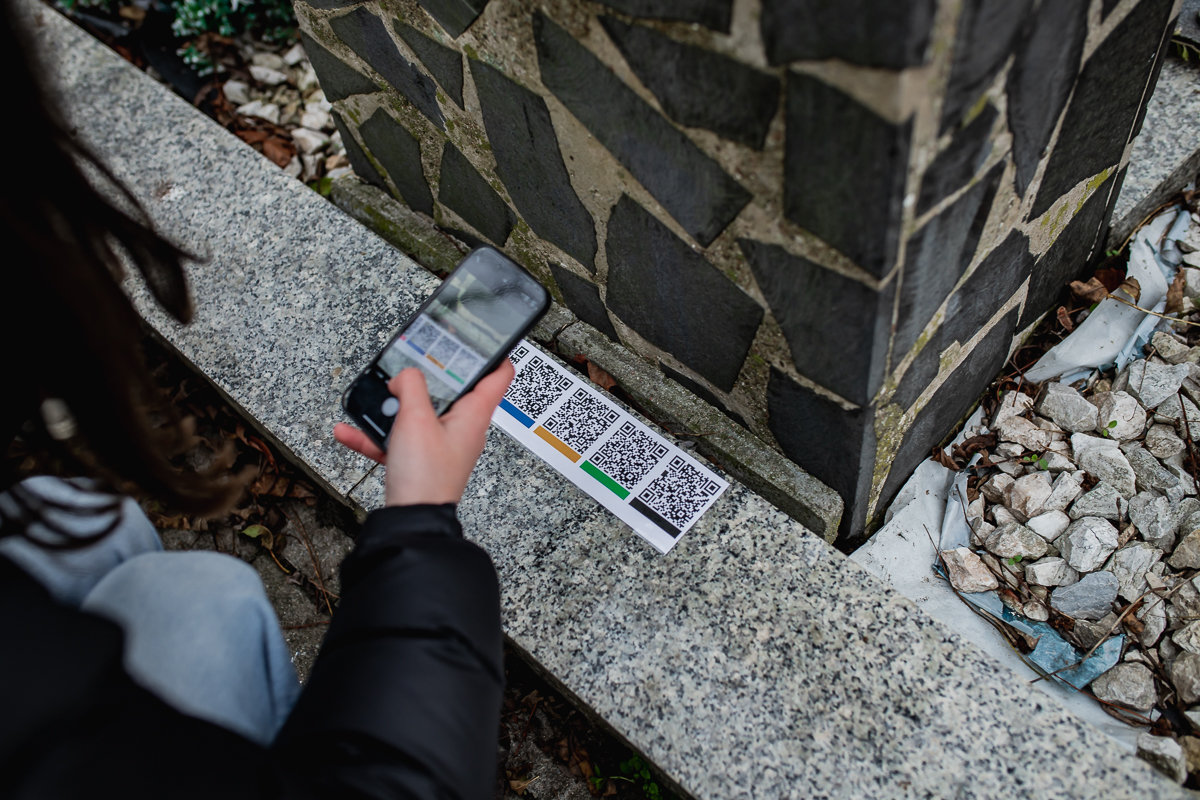 Na zdjęciu znajduje się kucająca na zewnątrz dziewczyna, trzymająca w dłoni smartfon i robiąca zdjęcie kodom QR umieszczonym w plenerze. Scena wygląda na element jakiejś gry terenowej lub zadania do wykonania na świeżym powietrzu.