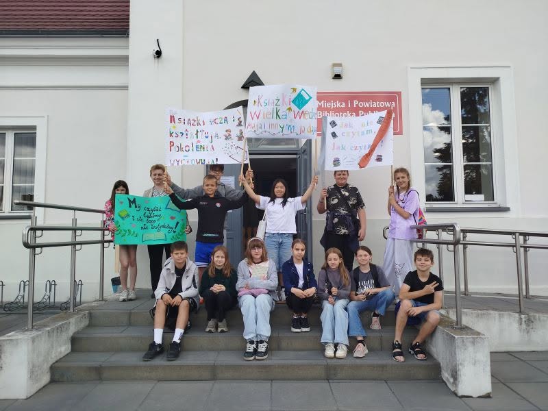 Grupa dzieci i dorosłych pozuje na schodach przed Miejską i Powiatową Biblioteką Publiczną. Uczestnicy trzymają ręcznie wykonane plakaty z hasłami promującymi czytelnictwo, m.in. Książki uczą i kształtują pokolenia oraz Książki Wielką Wiedzę dają.