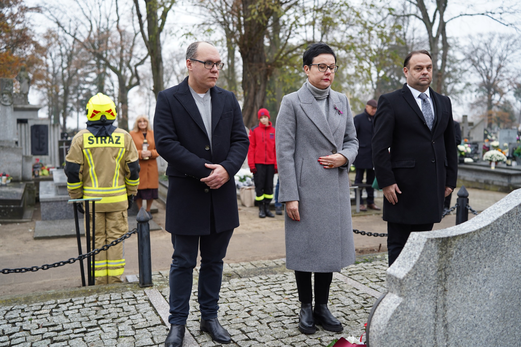 Na zdjęciu widać trzy osoby stojące przed nagrobkiem na cmentarzu, podczas uroczystości upamiętniającej. W tle stoi strażak w mundurze z napisem STRAŻ, kilka innych osób oraz groby ozdobione kwiatami. 