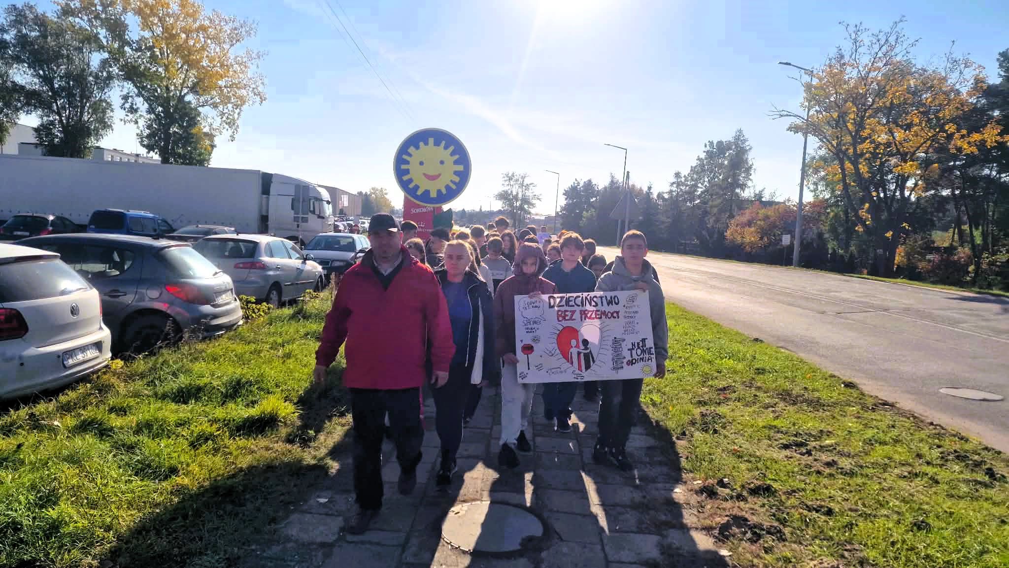 Uczniowie idą chodnikiem wzdłuż ulicy w słoneczny jesienny dzień. Na czele idzie mężczyzna w czerwonej kurtce. Młodzież niesie ręcznie malowany transparent z napisem Dzieciństwo bez przemocy i z sercem. W tle widać parking i żółte drzewa.