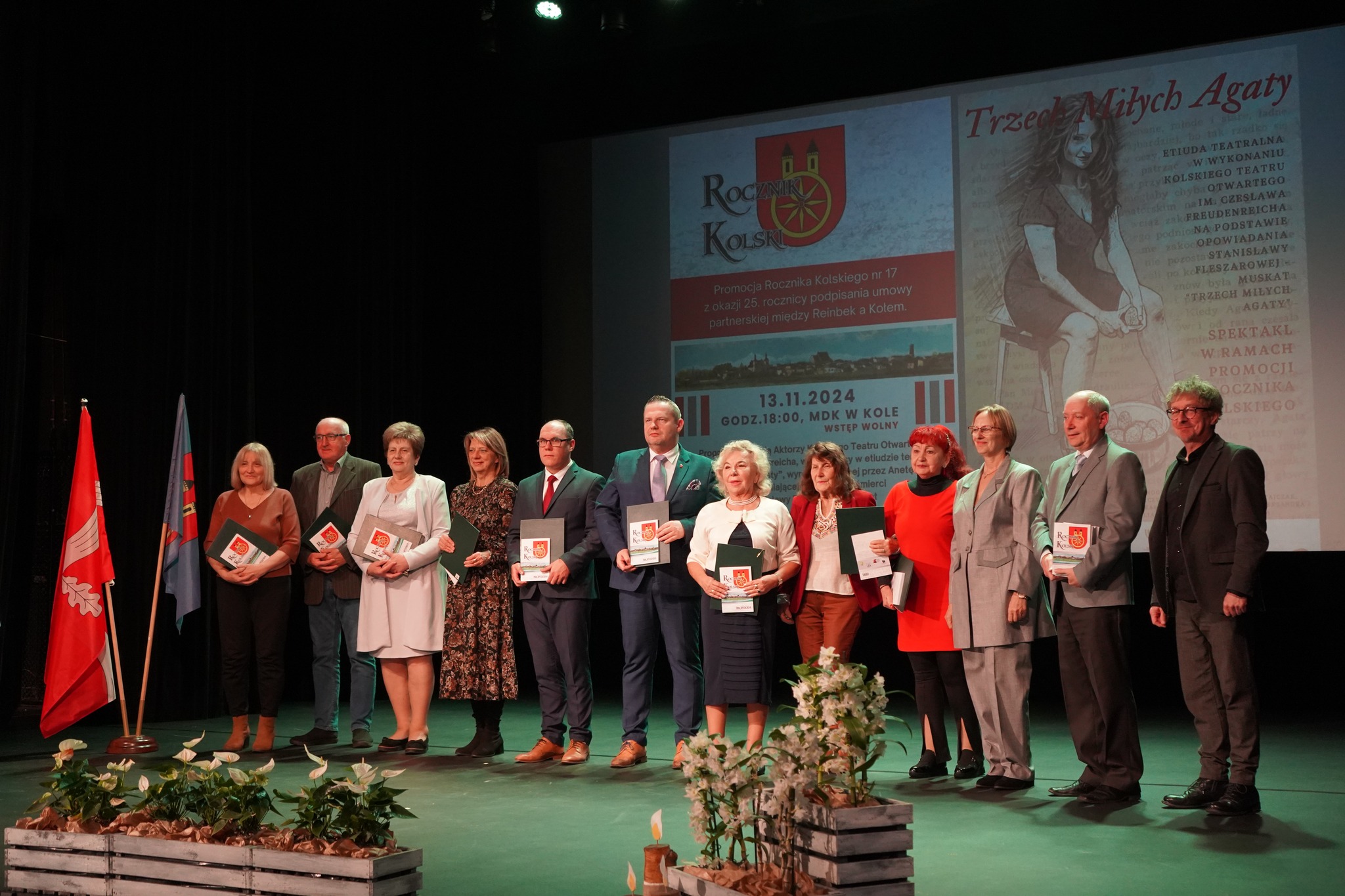 Grupa osób stoi na scenie, trzymając w rękach książki lub teczki, a w tle widnieje plakat promujący wydarzenie kulturalne, promocję Rocznika Kolskiego