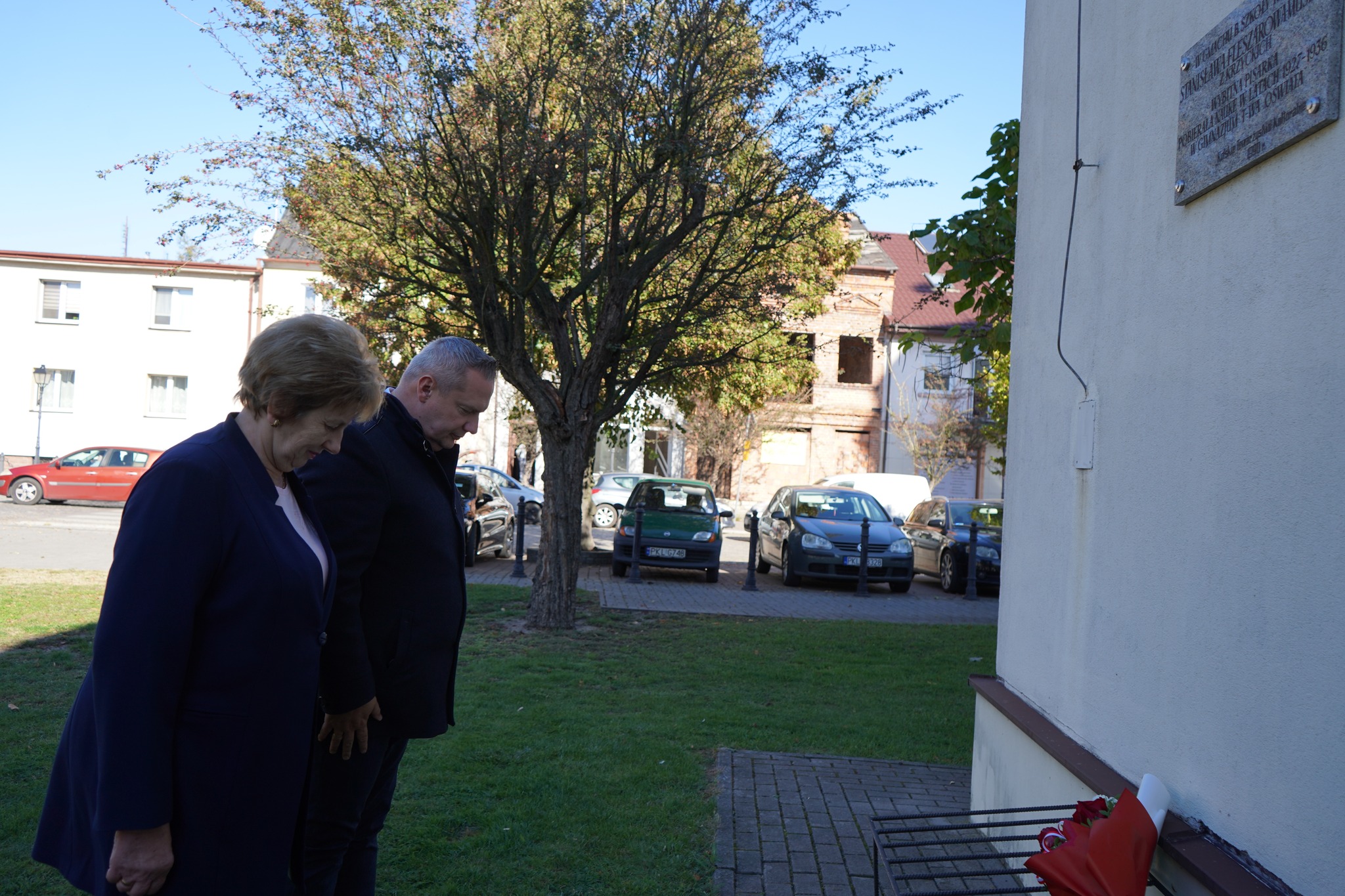 Na zdjęciu są dwie osoby – kobieta i mężczyzna, którzy pochylając głowy oddają hołd przed tablicą pamiątkową Stanisławy Fleszarowej – Muskat usytuowanej na ścianie Ratusza w Kole. W tle znajdują się pojazdy samochodowe i zabudowania kolskiej starówki