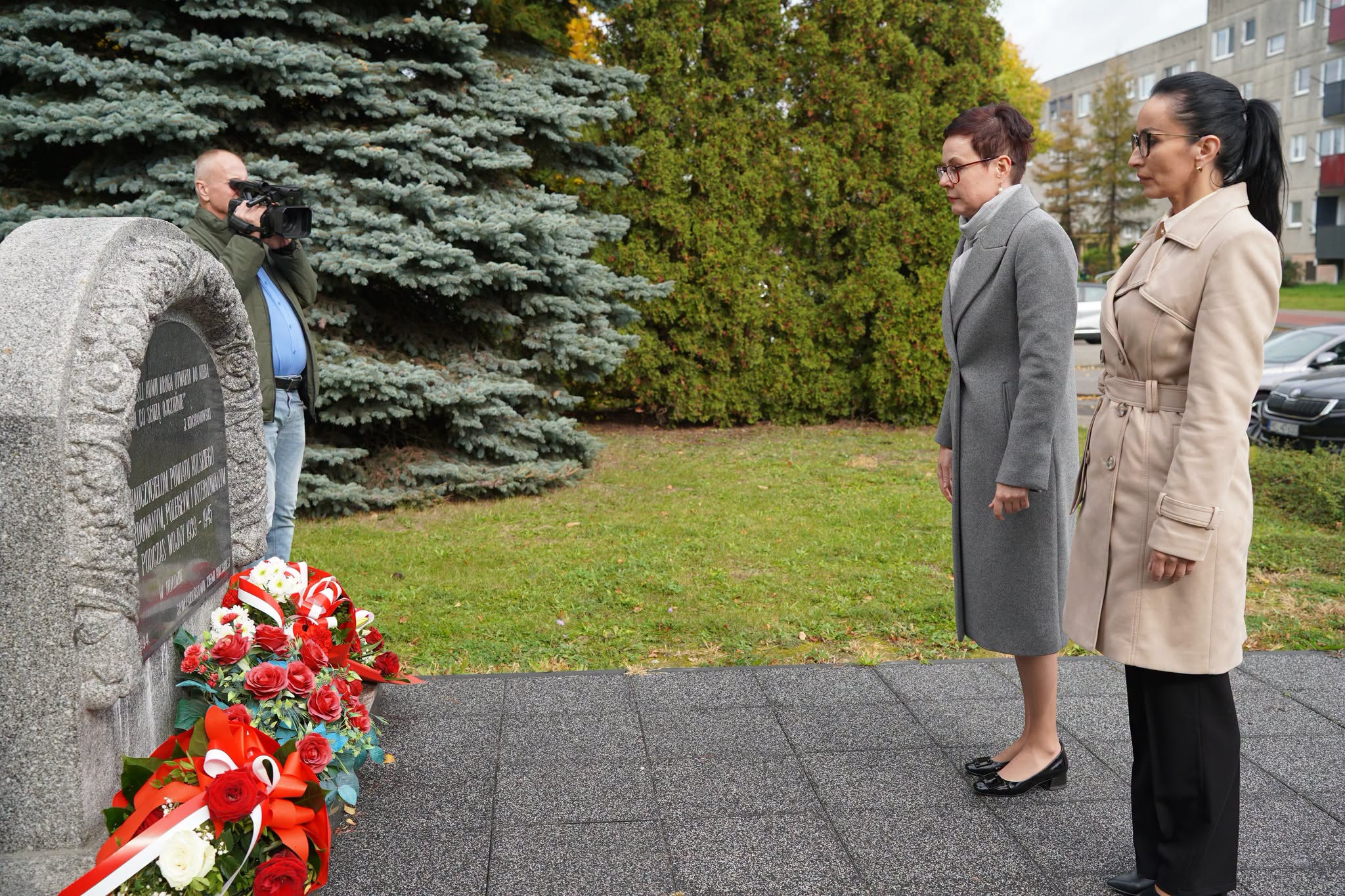Dwie kobiety w płaszczach, szarym i beżowym, stoją w zadumie przed granitowym pomnikiem z czarną tablicą, u którego podnóża leżą wieńce z czerwonych i białych kwiatów z biało-czerwonymi szarfami. Z boku operator z kamerą, w tle świerk i blok.