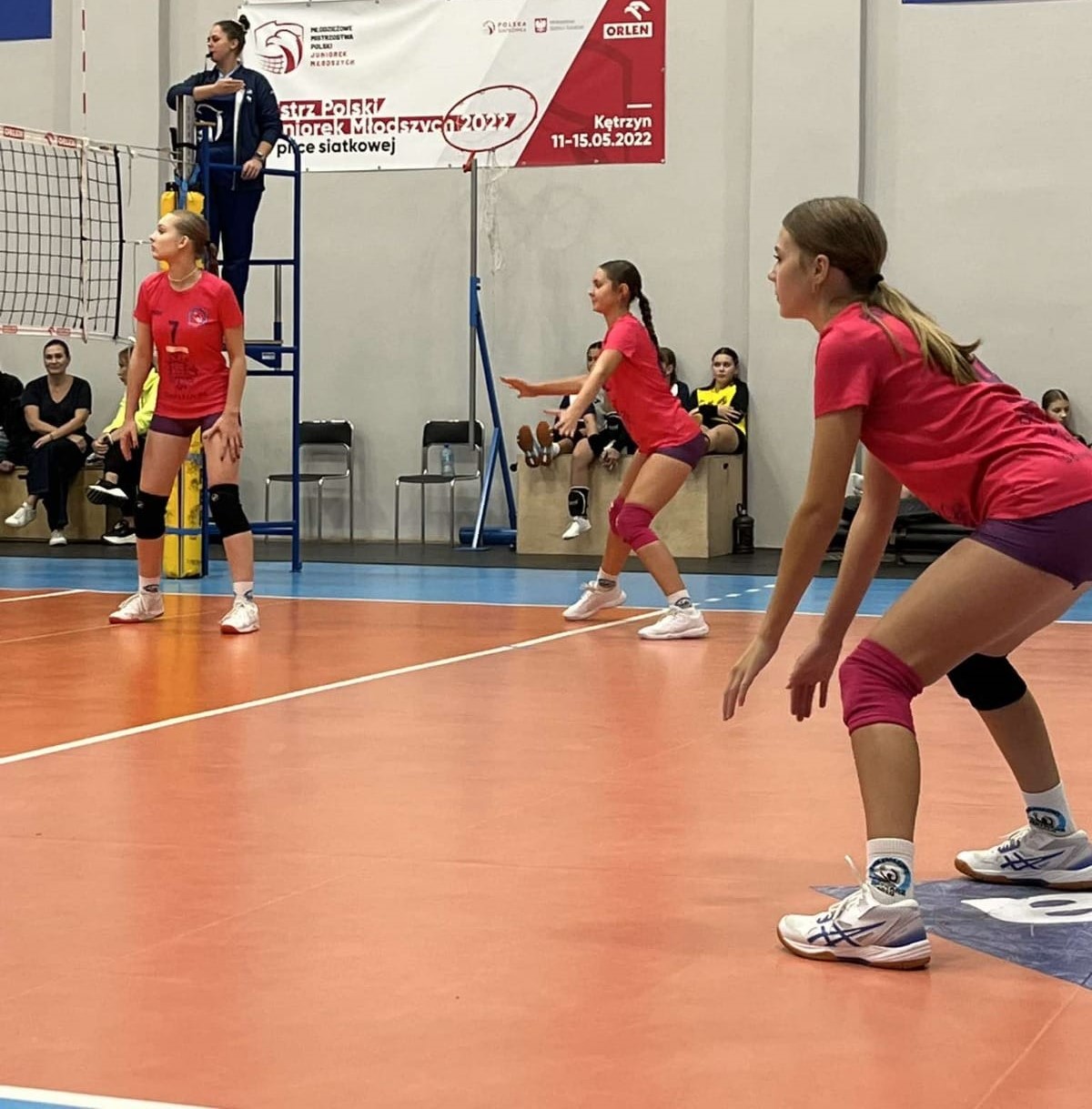 Na zdjęciu widoczne są trzy siatkarki w trakcie meczu siatkarskiego. Mecz rozgrywany jest w hali sportowej, w tle widać sędzinę z gwizdkiem w ustach z podniesioną prawą dłonią oraz wiszący banner reklamowy na ścianie.