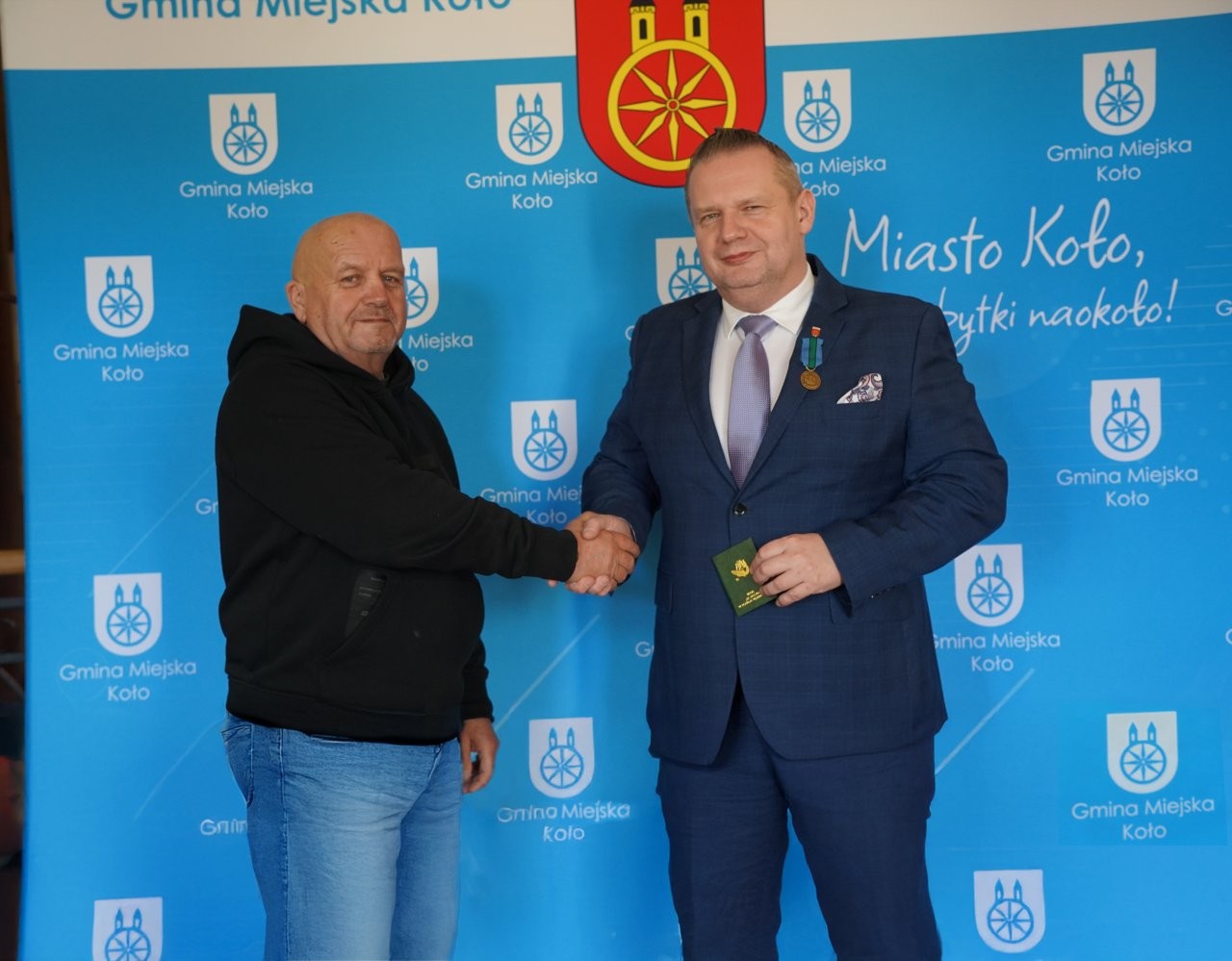 Na zdjęciu dwóch mężczyzn wita się, podając sobie obie dłonie w geście gratulacji. Mężczyzna po prawej ma na sobie garnitur z przypiętym odznaczeniem i trzyma w rękach niewielką książeczkę wędkarską. W tle znajduje się ścianka promocyjna miasta.