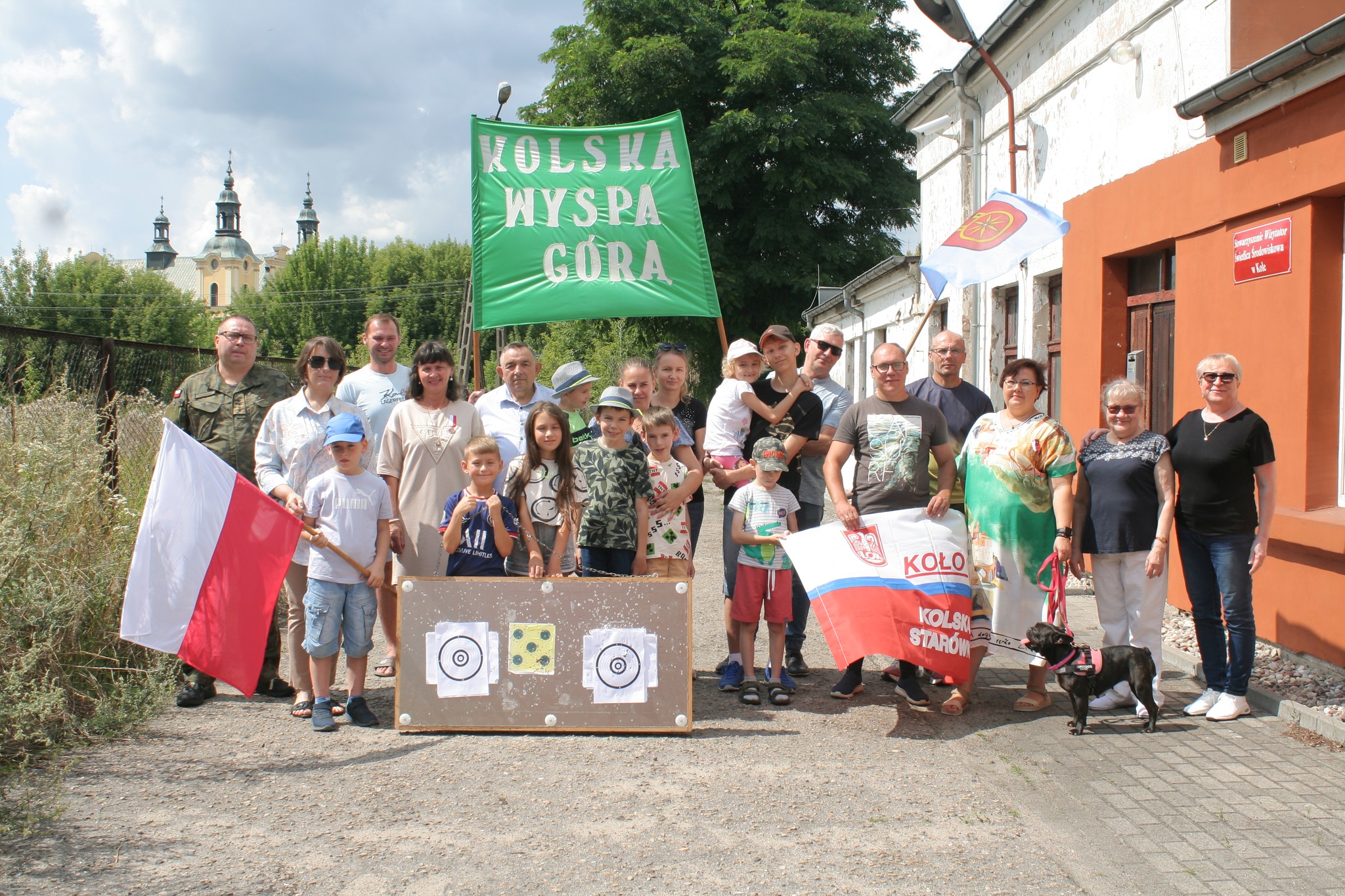 Na zdjęciu widoczna jest grupa ludzi – dzieci i dorośli – zgromadzona przed budynkiem. Trzymają flagi Polski, Koła oraz zielony transparent z napisem „Kolska Wyspa Góra”. Przed nimi stoi tablica z tarczami strzelniczymi.