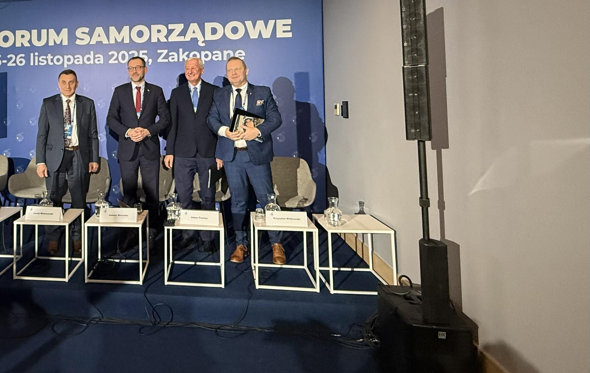 Na zdjęciu widać czterech mężczyzn w garniturach stojących na podwyższonym podeście, będącym częścią panelu dyskusyjnego. W tle znajduje się baner z napisem „Forum Samorządowe”. Przed nimi stoją małe stoliczki, a za nimi ustawiono fotele.