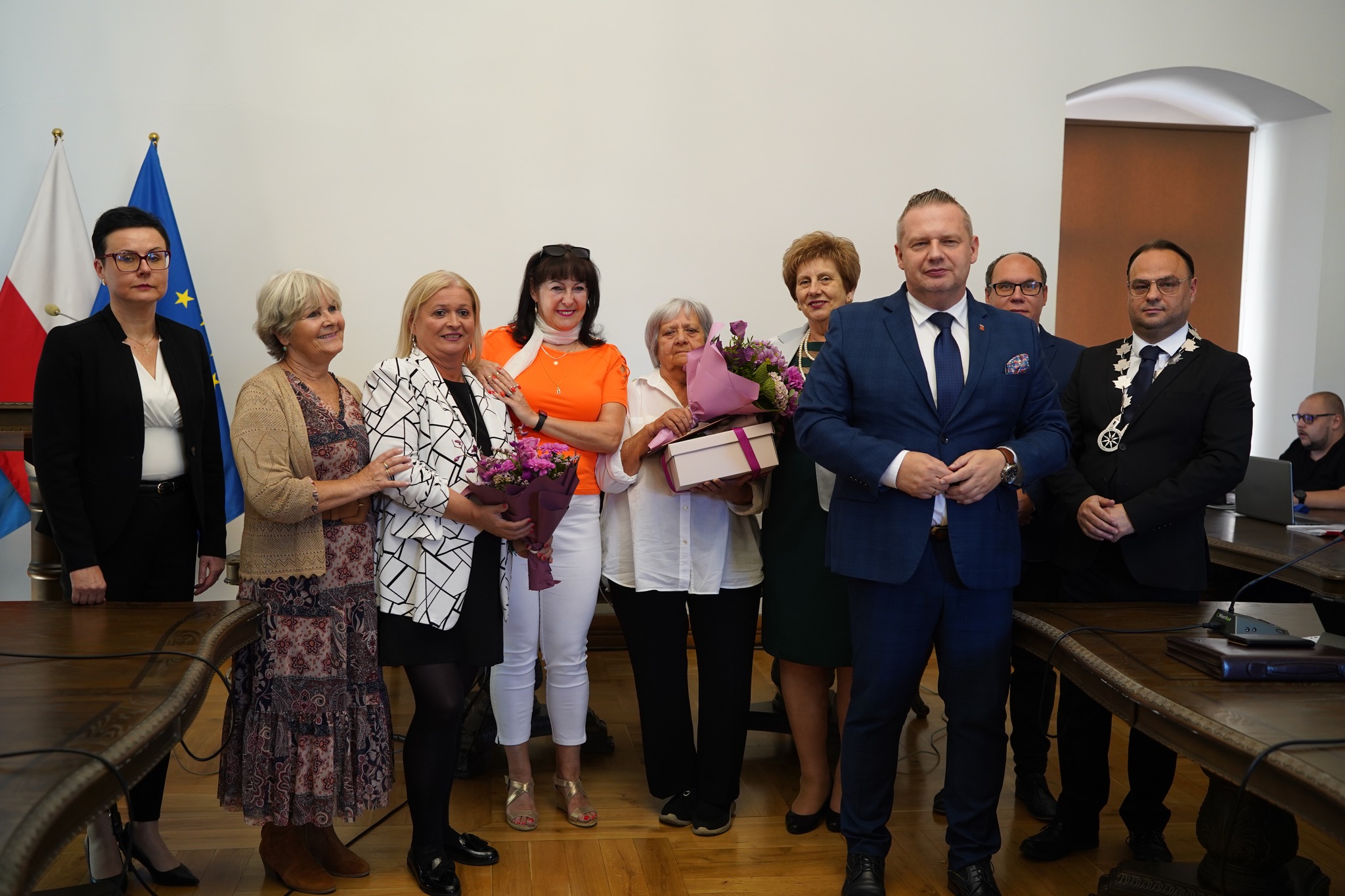 Na zdjęciu widać grupę osób podczas oficjalnego spotkania – kilka kobiet trzyma bukiety kwiatów i upominek, a obok nich stoją elegancko ubrani mężczyźni i kobiety. W tle flagi Polski i UE. Jest to uroczystość wręczenia gratulacji.