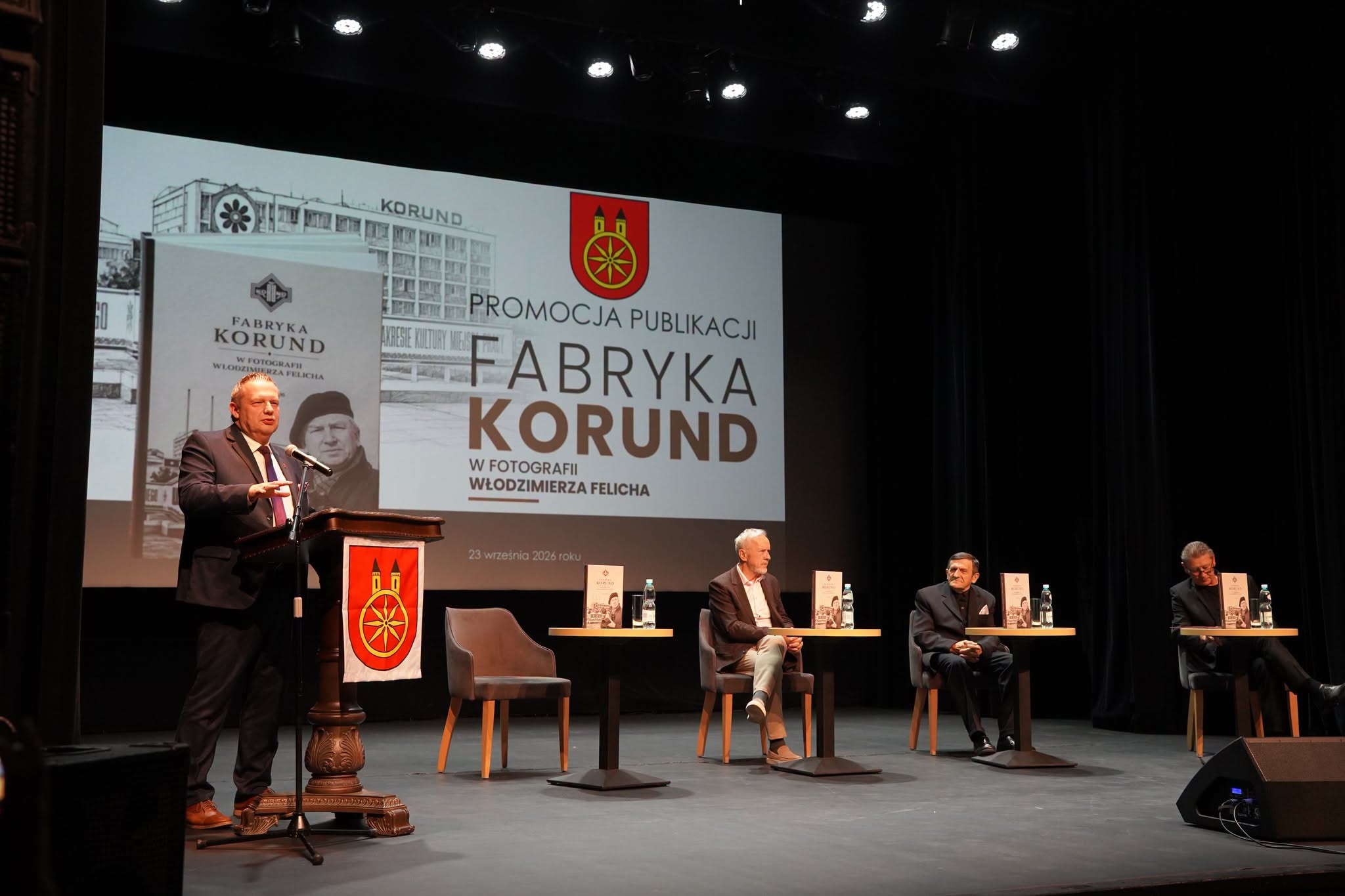 Mężczyzna przemawia przy mównicy z herbem miasta na tle ekranu z napisem Promocja publikacji Fabryka Korund w fotografii Włodzimierza Felicha. Obok scena z trzema siedzącymi mężczyznami i stolikami z egzemplarzami książki.