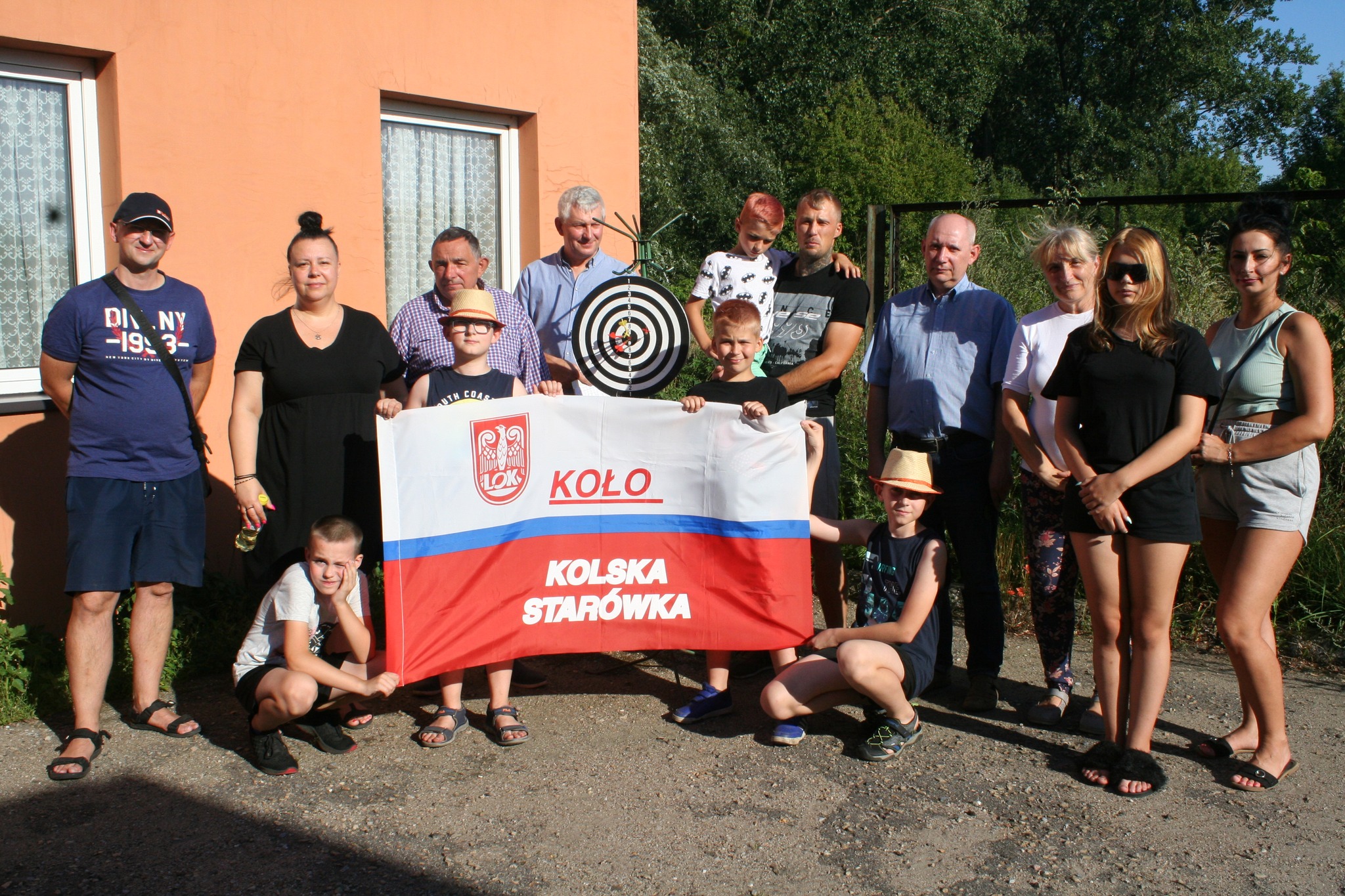 Na zdjęciu znajduje się grupa ludzi stojąca na zewnątrz przed budynkiem z pomarańczową elewacją. Uczestnicy pozują do zdjęcia z flagą i tarczą strzelniczą. Na fladze widnieje napis KOŁO KOLSKA STARÓWKA oraz logo organizacji LOK (Liga Obrony Kraju)