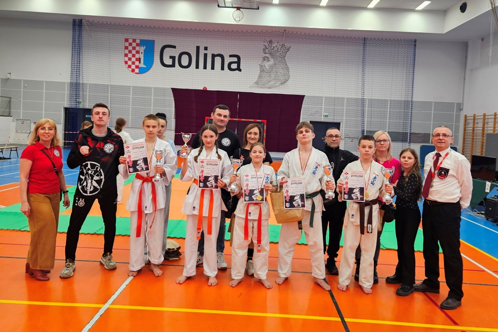Grupa karateków, dziewcząt i chłopców wraz z trenerami, w hali sportowej, pozuje do zdjęcia z pucharami i dyplomami na Otwartych Mistrzostwach o Puchar Burmistrza Goliny. W tle widać bramkę oraz herb i napis GOLINA.