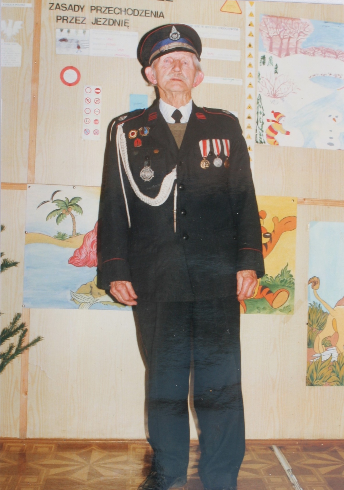 Na zdjęciu widać starszego mężczyznę w galowym mundurze strażackim z medalami, stojącego na tle dziecięcych rysunków i plakatów.