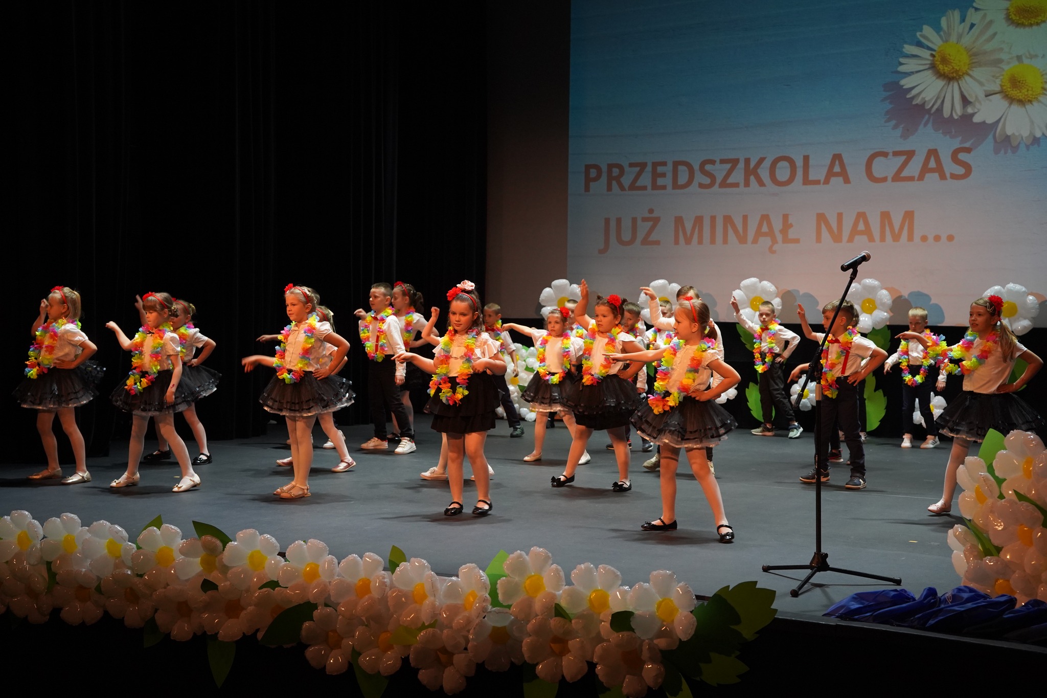 Na zdjęciu widać grupę dzieci występujących na scenie podczas uroczystości zakończenia przedszkola. Dzieci są kolorowo ubrane, a tło zdobi napis „Przedszkola czas już minął nam…”.
