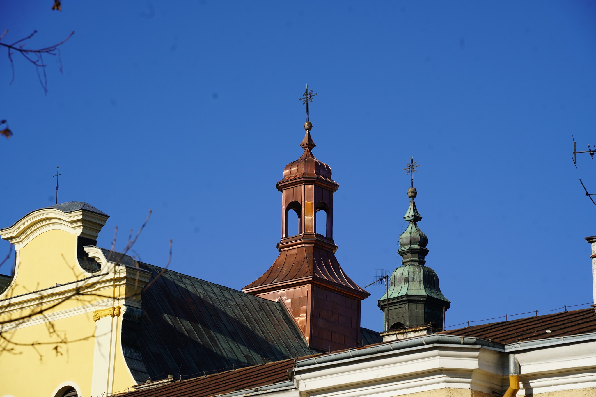 Na zdjęciu widać fragment dachu zabytkowego klasztoru. Widoczne są dwie sygnaturki z krzyżami na szczytach - miedziana wieżyczka o ciepłym odcieniu oraz zielonkawa, patynowana kopuła. Całość znajduje się na tle czystego, błękitnego nieba.