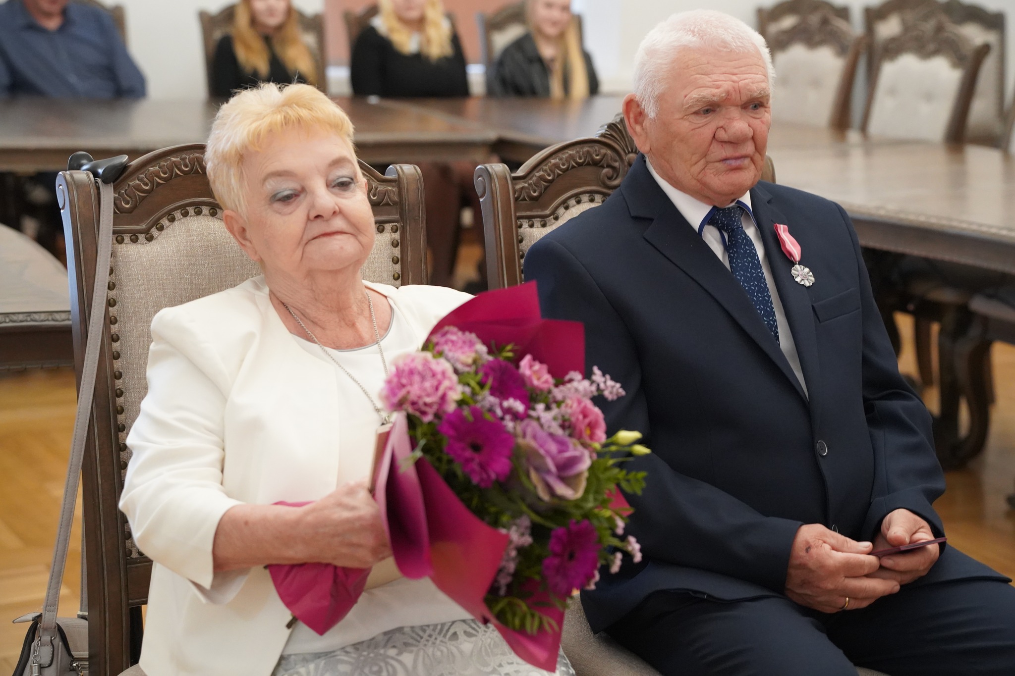 Na zdjęciu starsze małżeństwo siedzi obok siebie podczas uroczystości jubileuszu Złotych Godów - kobieta trzyma bukiet kwiatów, a mężczyzna ma medal przypięty do marynarki