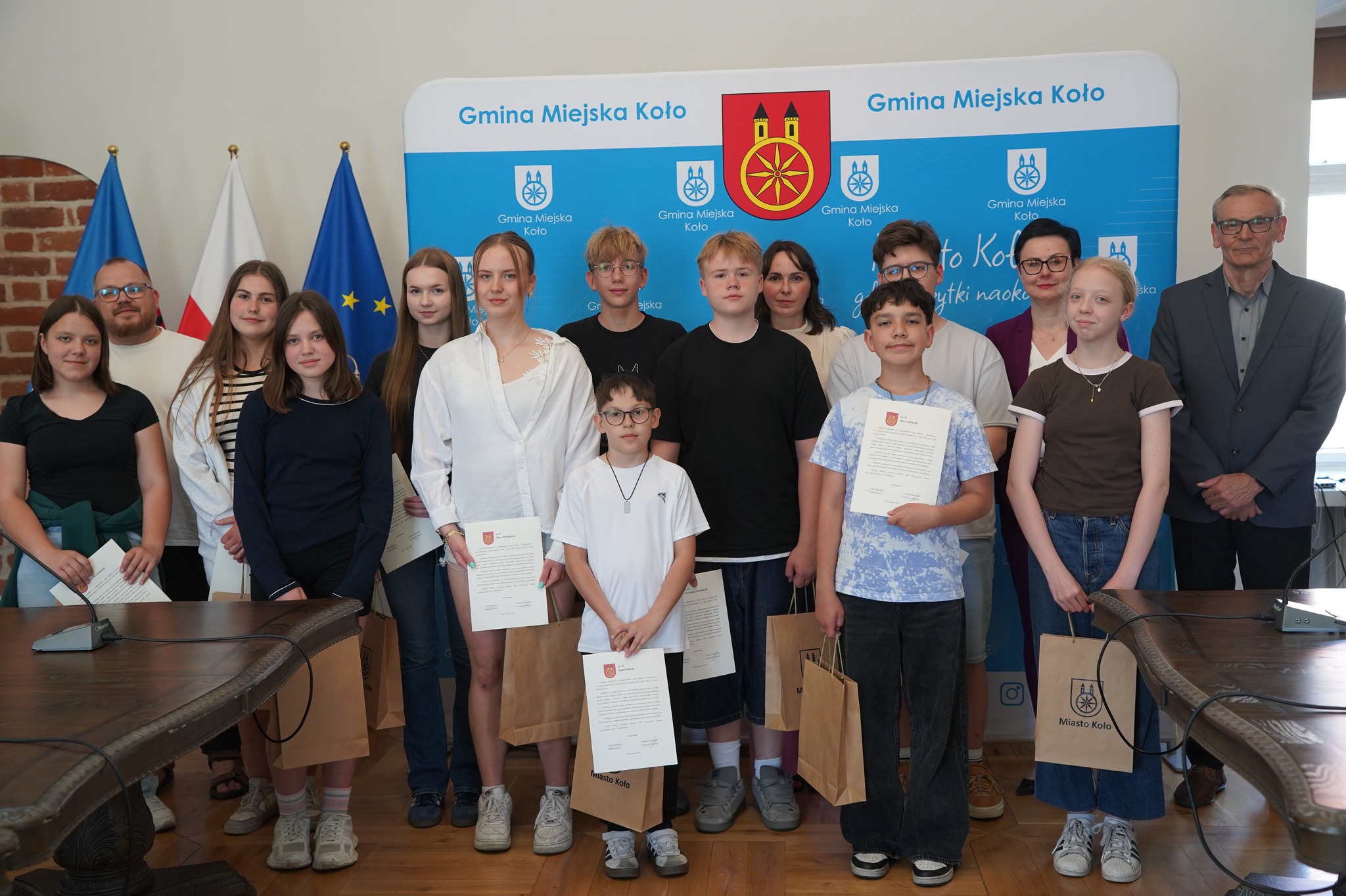 Grupa uczniów i dorosłych pozuje do zdjęcia w sali urzędu na tle baneru Gmina Miejska Koło. Dzieci trzymają dyplomy i papierowe torby z logo Miasto Koło. W tle flagi Polski i Unii Europejskiej.
