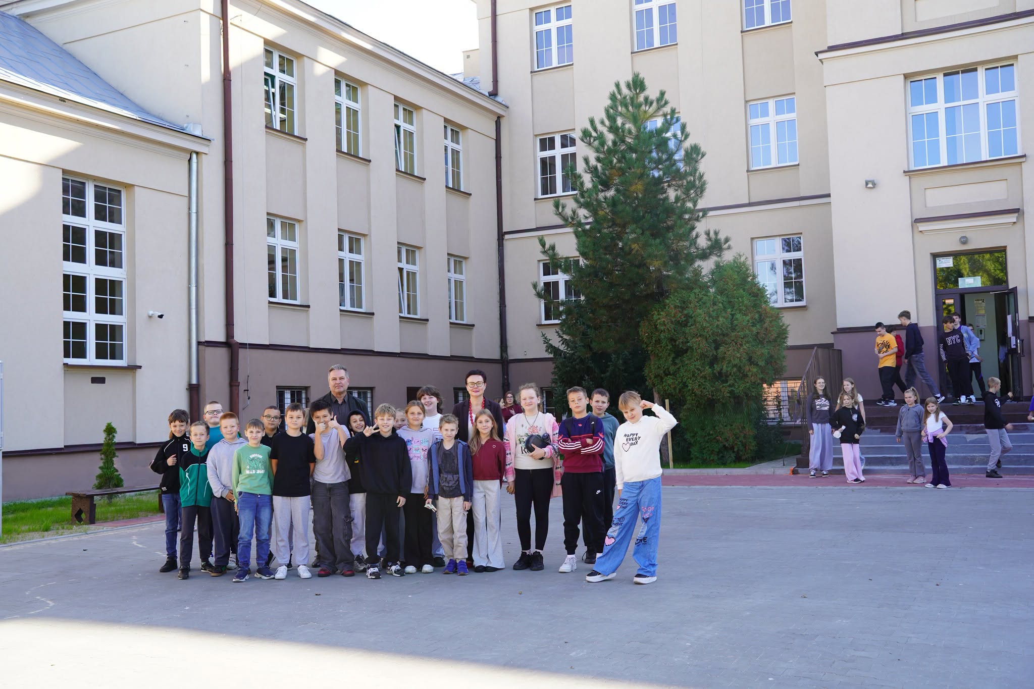 Grupa dzieci i dwóch dorosłych pozuje do zdjęcia na dziedzińcu szkolnym przed beżowym budynkiem szkoły. W tle inni uczniowie wychodzący z budynku, zielone drzewo i schody wejściowe.