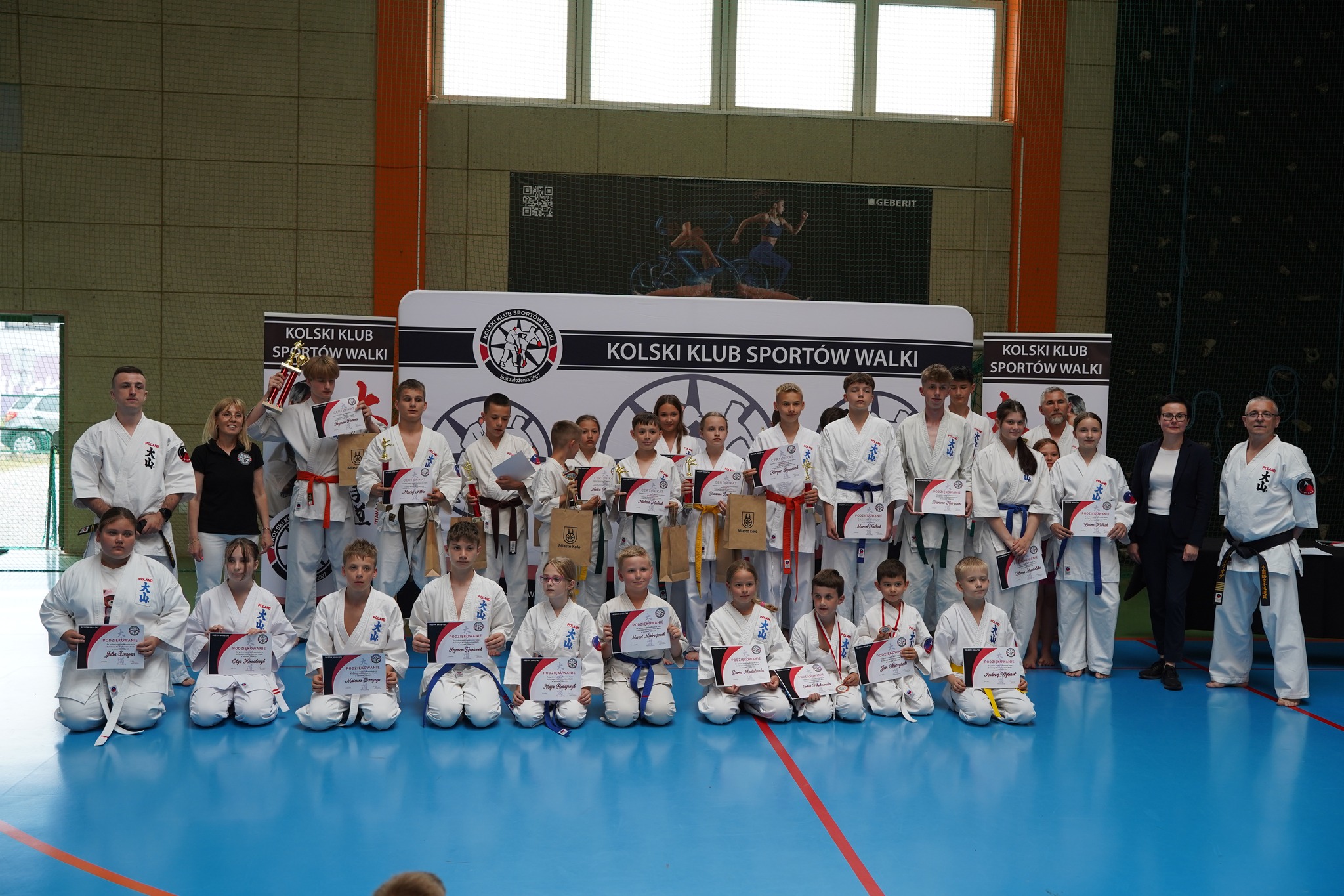 Na zdjęciu widać grupę uczestników turnieju sztuk walki zorganizowanego przez Kolski Klub Sportów Walki — dzieci i młodzież w kimonach prezentują dyplomy i trofea.