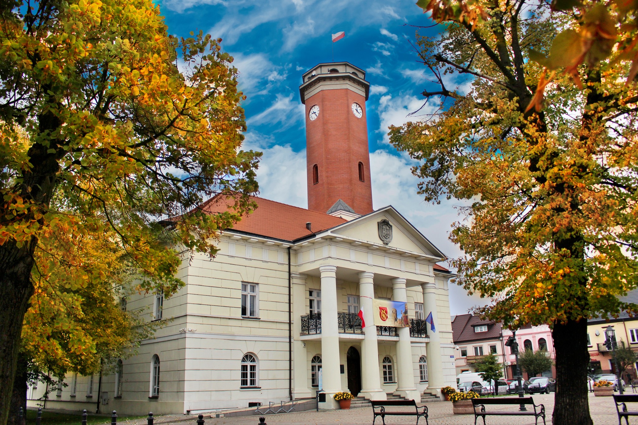 Zdjęcie przedstawia ratusz w mieście Koło. Budynek ma klasyczny wygląd, z charakterystycznymi białymi kolumnami na fasadzie, które wspierają balkon nad wejściem. Widać również czerwoną wieżę zegarowa z polską flagą na szczycie. Ratusz otaczają drzewa