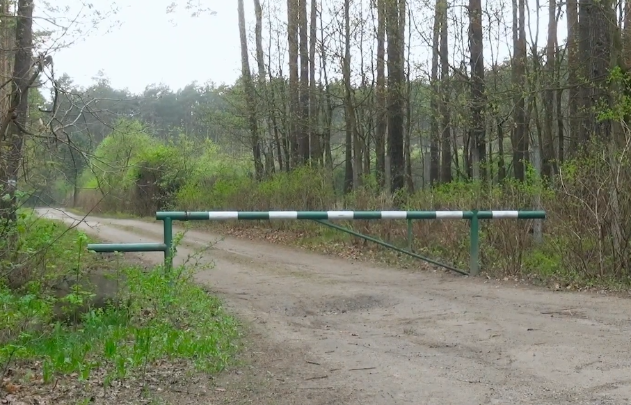 Na zdjęciu widoczny jest szlaban ograniczający wjazd do lasu. W tle rozciąga się leśna sceneria pełna drzew i zieleni, podkreślająca spokojny oraz naturalny charakter otoczenia.