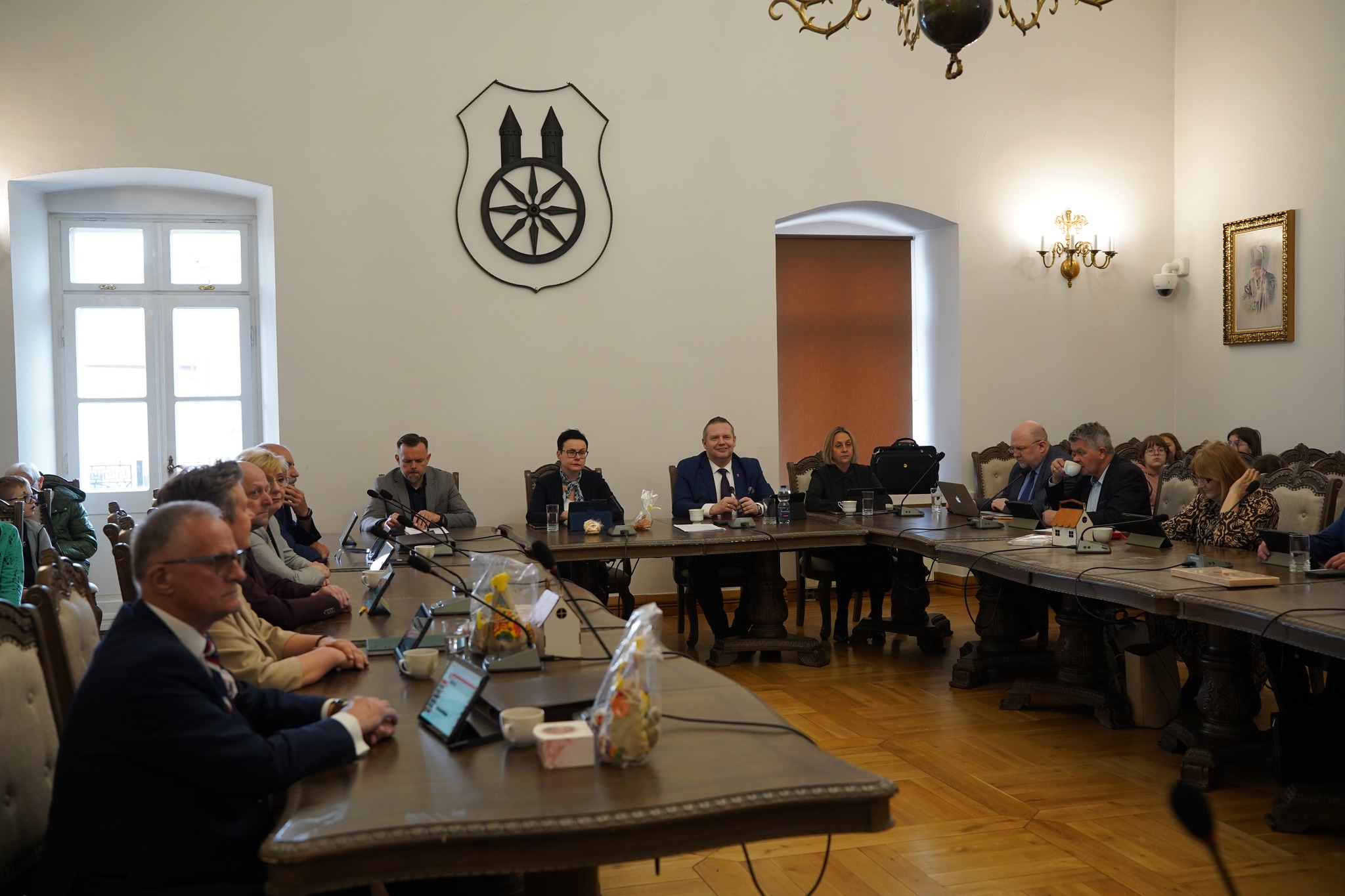 Na zdjęciu widać salę obrad, w której przy dużych stołach siedzą uczestnicy oficjalnego posiedzenia. Osoby mają przed sobą dokumenty i tablety, a całość ma charakter formalnego spotkania samorządowego.