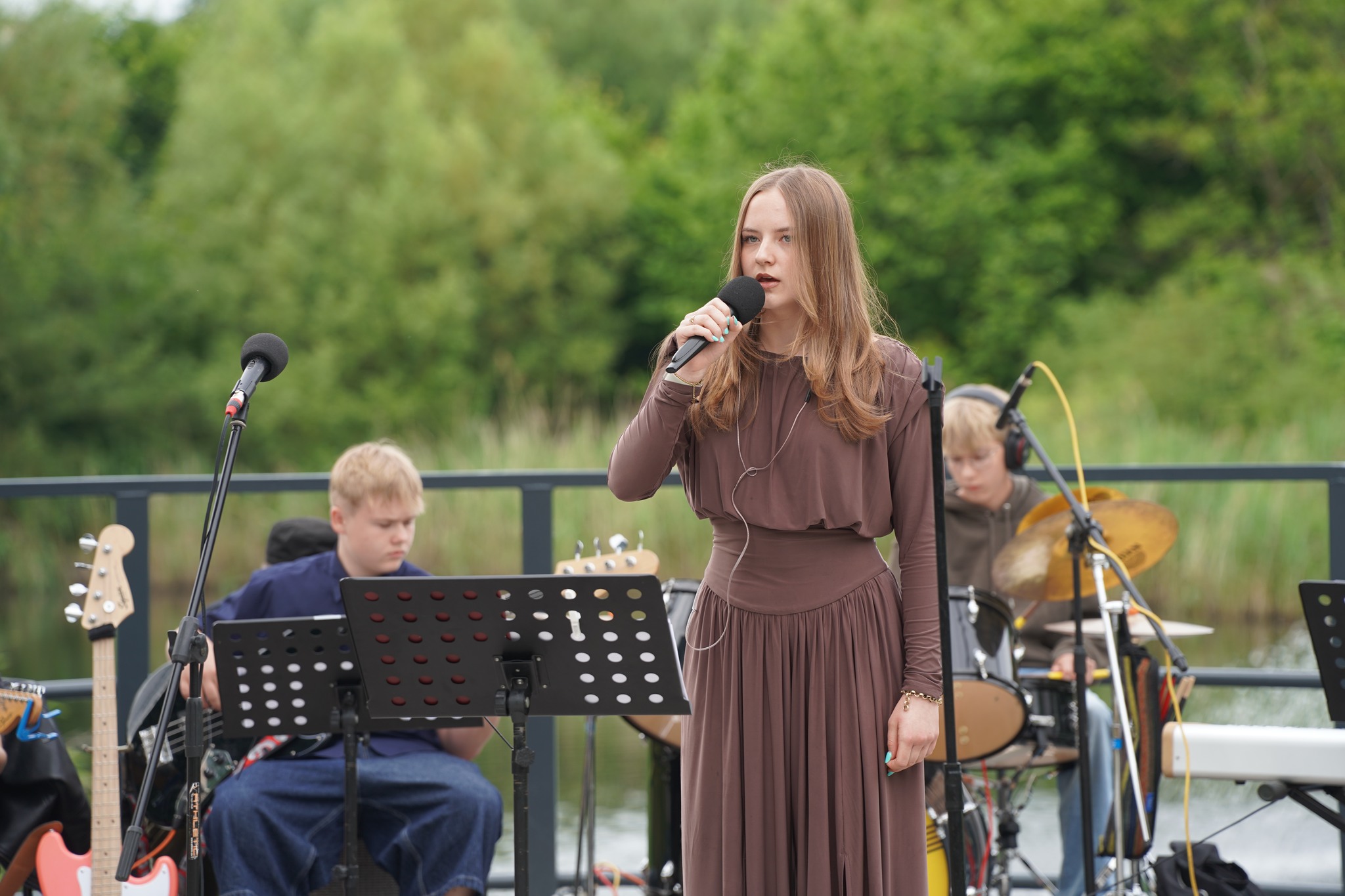 Młoda wokalistka śpiewa do mikrofonu na scenie plenerowej nad wodą. W tle zespół muzyczny: gitarzysta i perkusista. 