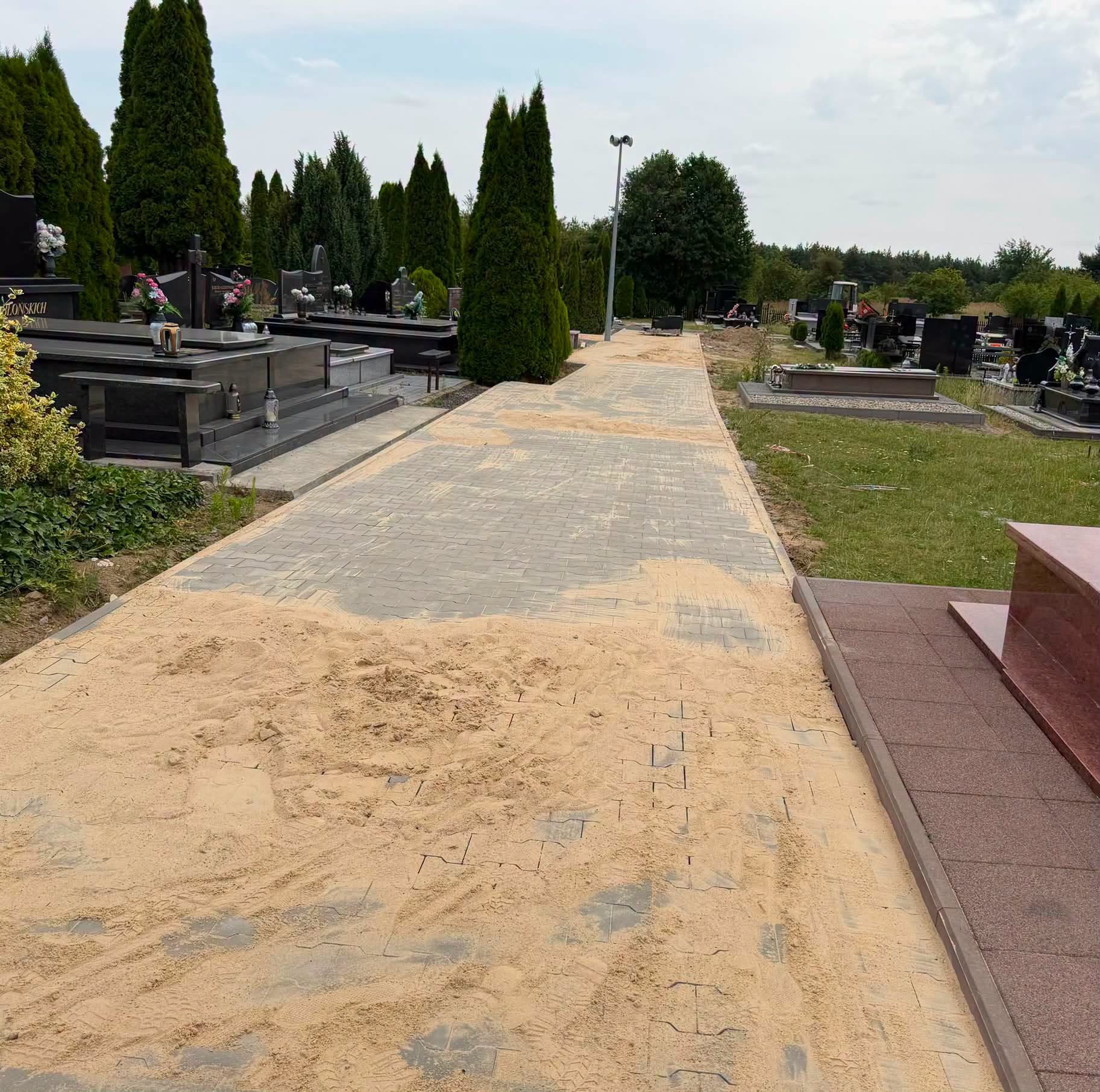 Alejka cmentarna wyłożona szarą kostką brukową, częściowo pokryta piaskiem w trakcie układania. Po obu stronach ciemne nagrobki i tuje, w tle las i słup oświetleniowy.