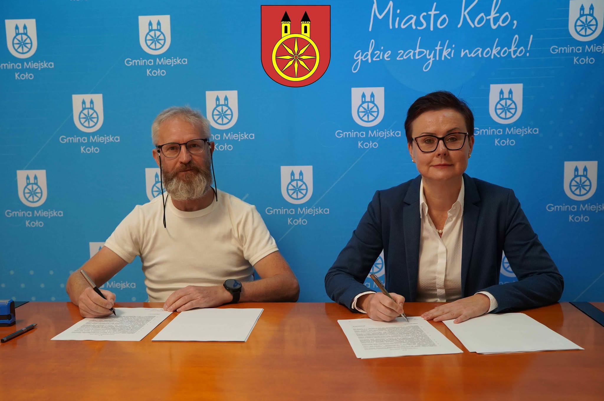 Dwie osoby siedzące przy stole (po prawej kobieta, a po lewej mężczyzna) podpisujące umowę. W tle widać ściankę reklamową Gminy Miejskiej Koło.
