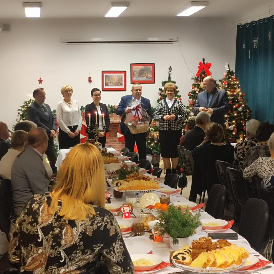 Na zdjęciu widać osoby siedzące przy wigilijnym stole z tradycyjnymi potrawami, zwrócone w stronę stojącej delegacji władz samorządowych. W tle znajdują się świątecznie ozdobione choinki, podkreślające bożonarodzeniową atmosferę.