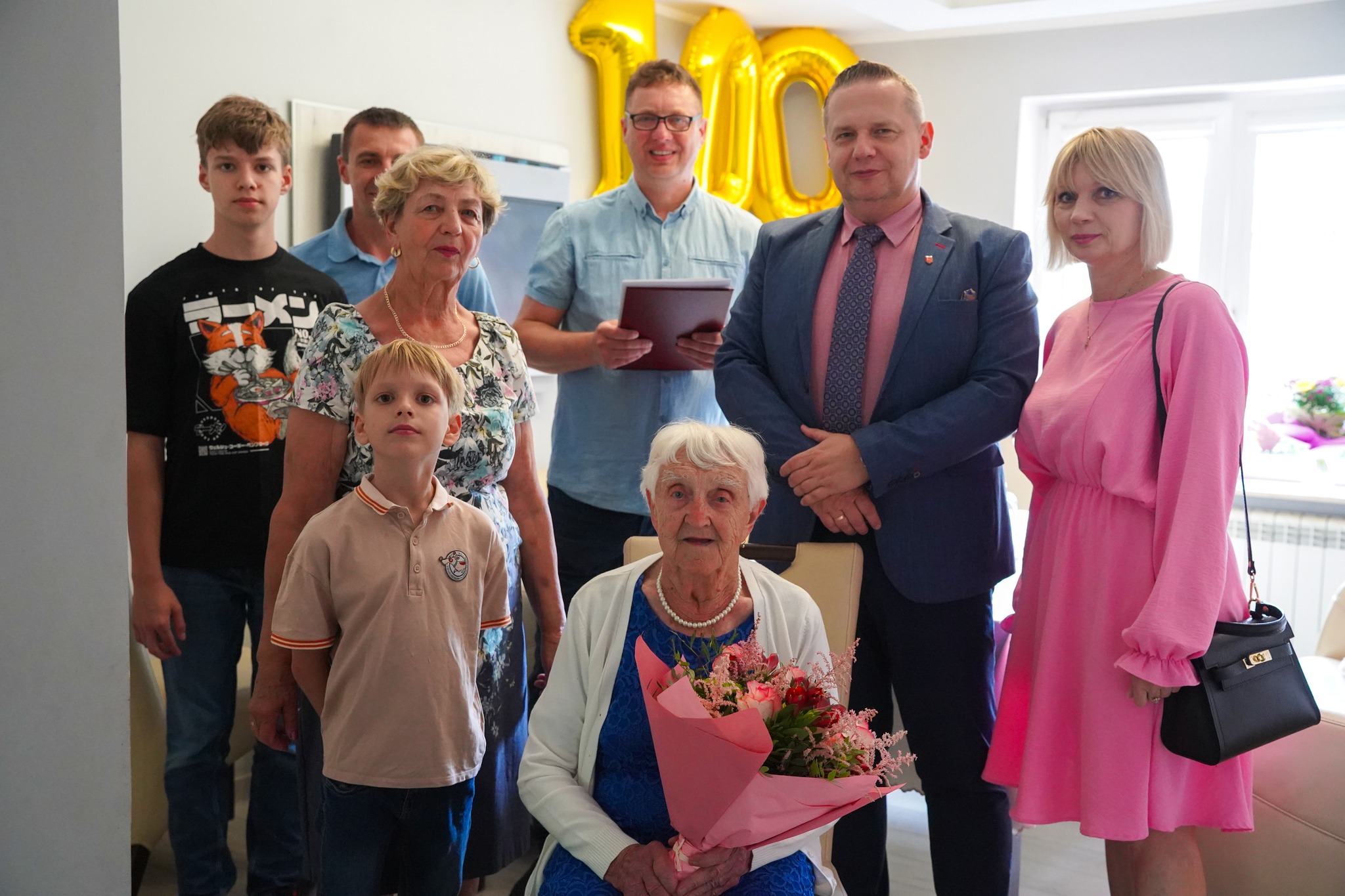 Na zdjęciu widać starszą kobietę obchodzącą 100. urodziny, otoczoną przez rodzinę i bliskich oraz przedstawicieli władz samorządowych miasta Koła. Trzyma bukiet kwiatów, a w tle znajdują się złote balony tworzące liczbę „100”.